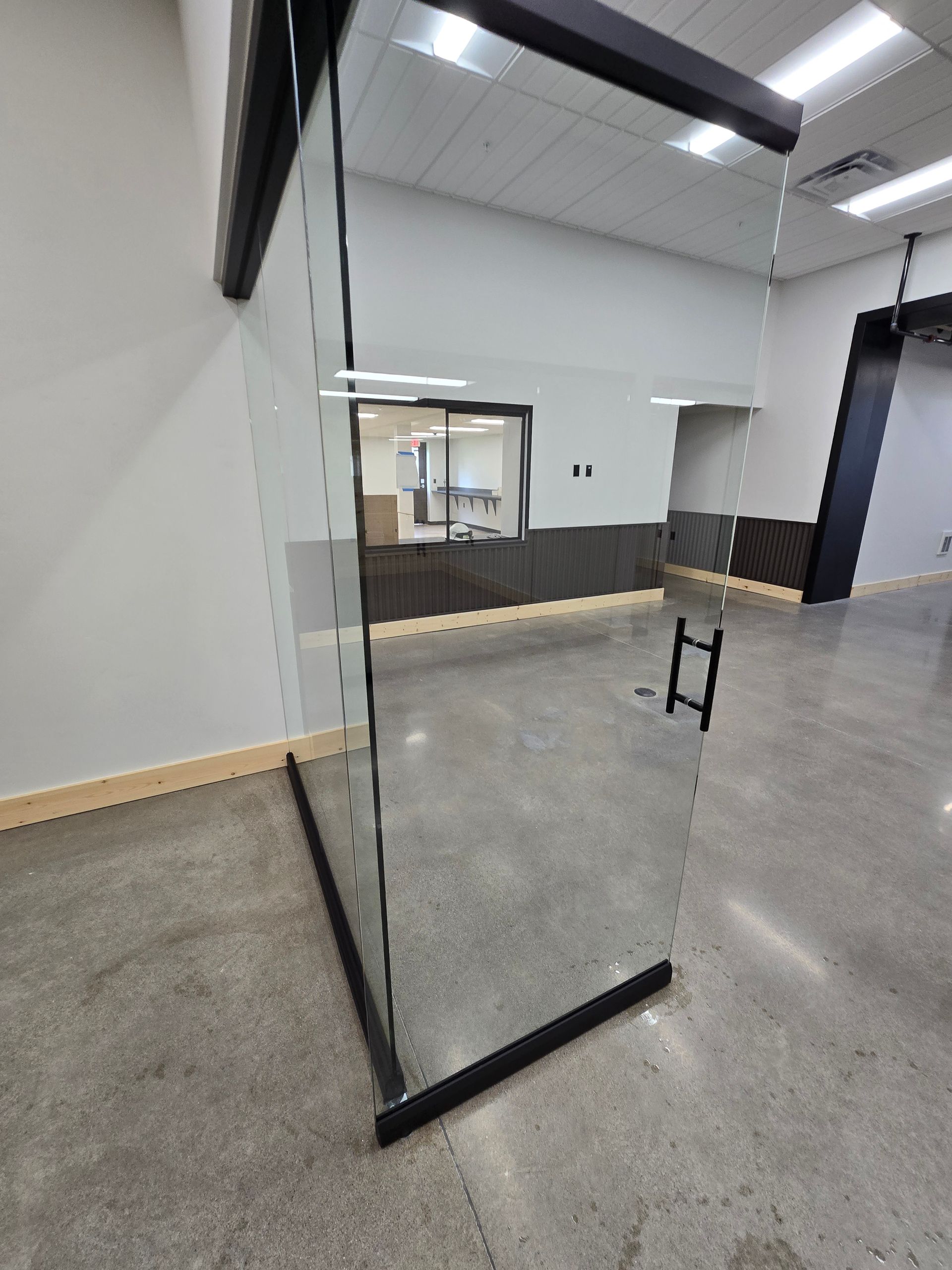 A glass door with a black handle and frame in a modern office hallway with concrete floors.