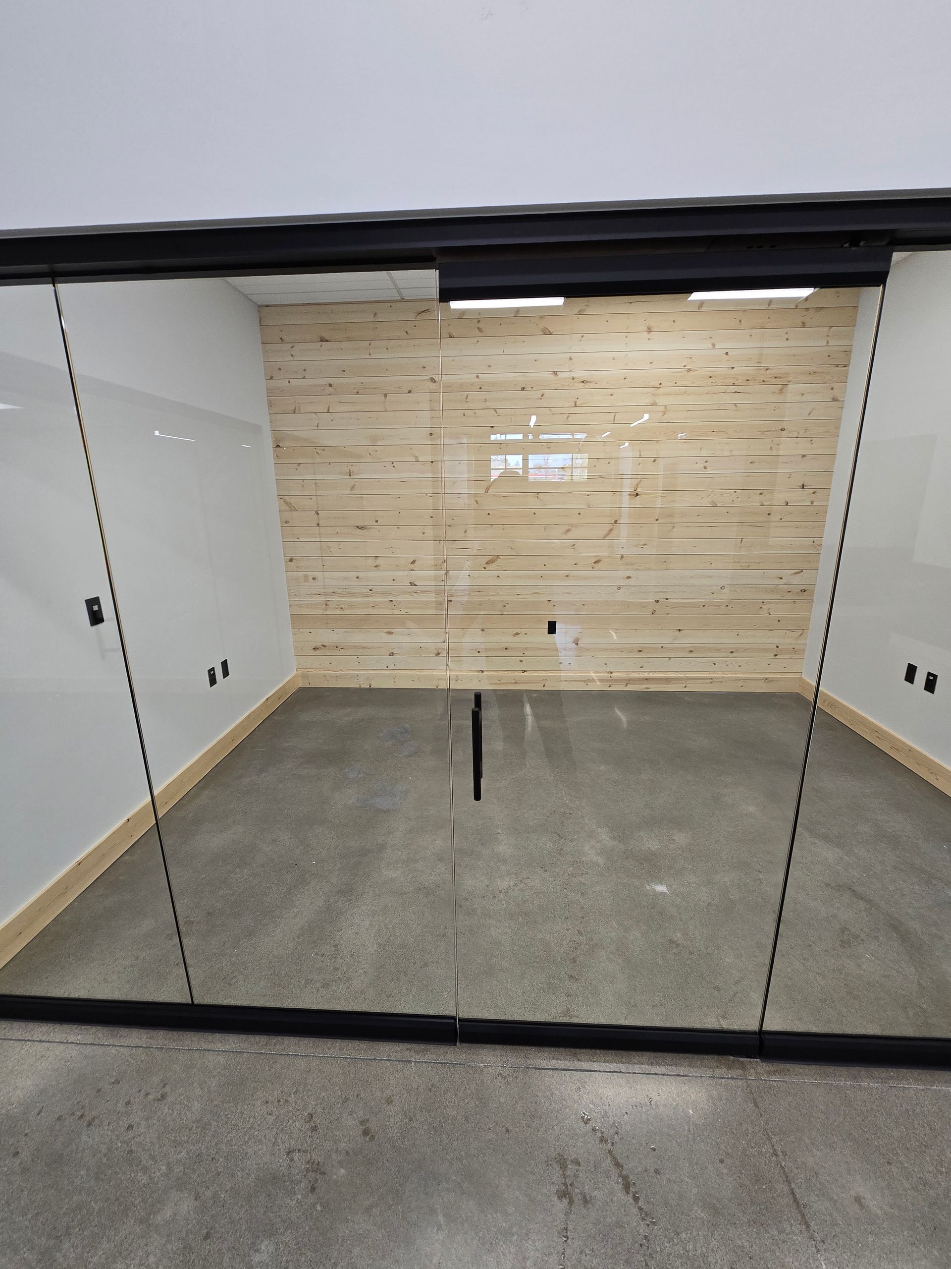A glass-walled office pod with a light-colored wood paneled back wall and grey concrete floors.