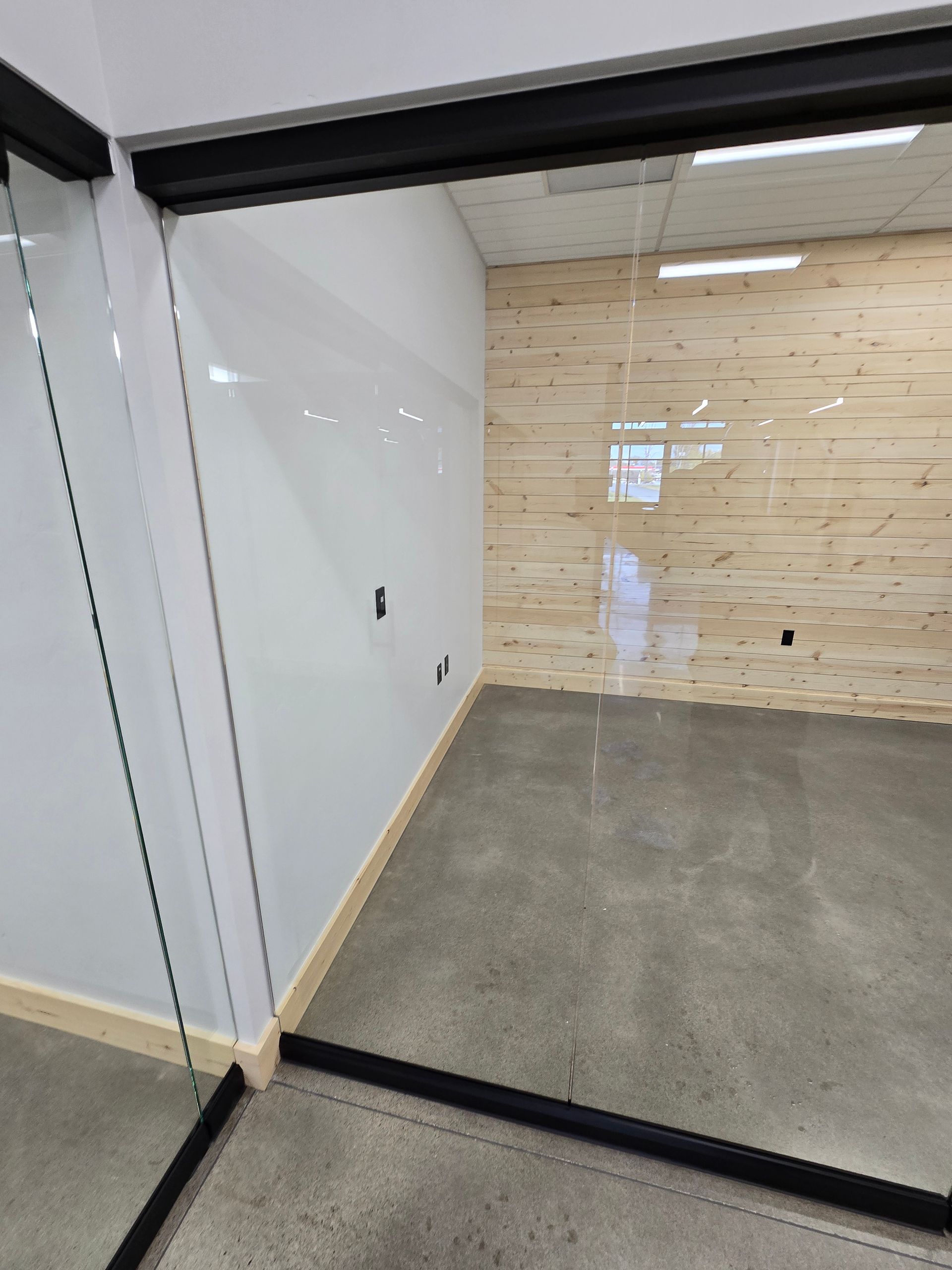 An empty office space with a glass wall, white side wall, wood-paneled back wall, and polished concrete floor.