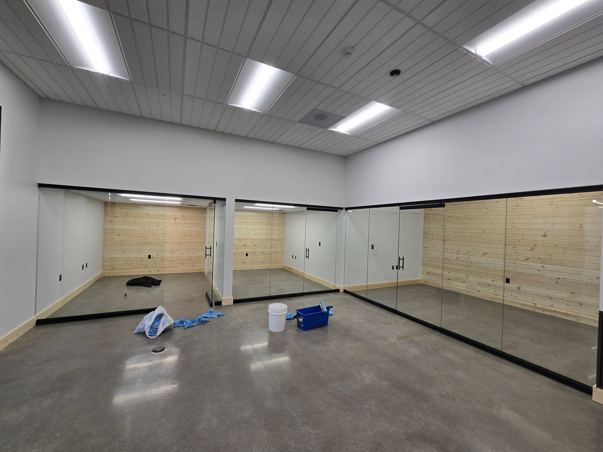 A dance or gym studio featuring floor-to-ceiling mirrors, light wood paneled walls, gray concrete floors, and ceiling lights.