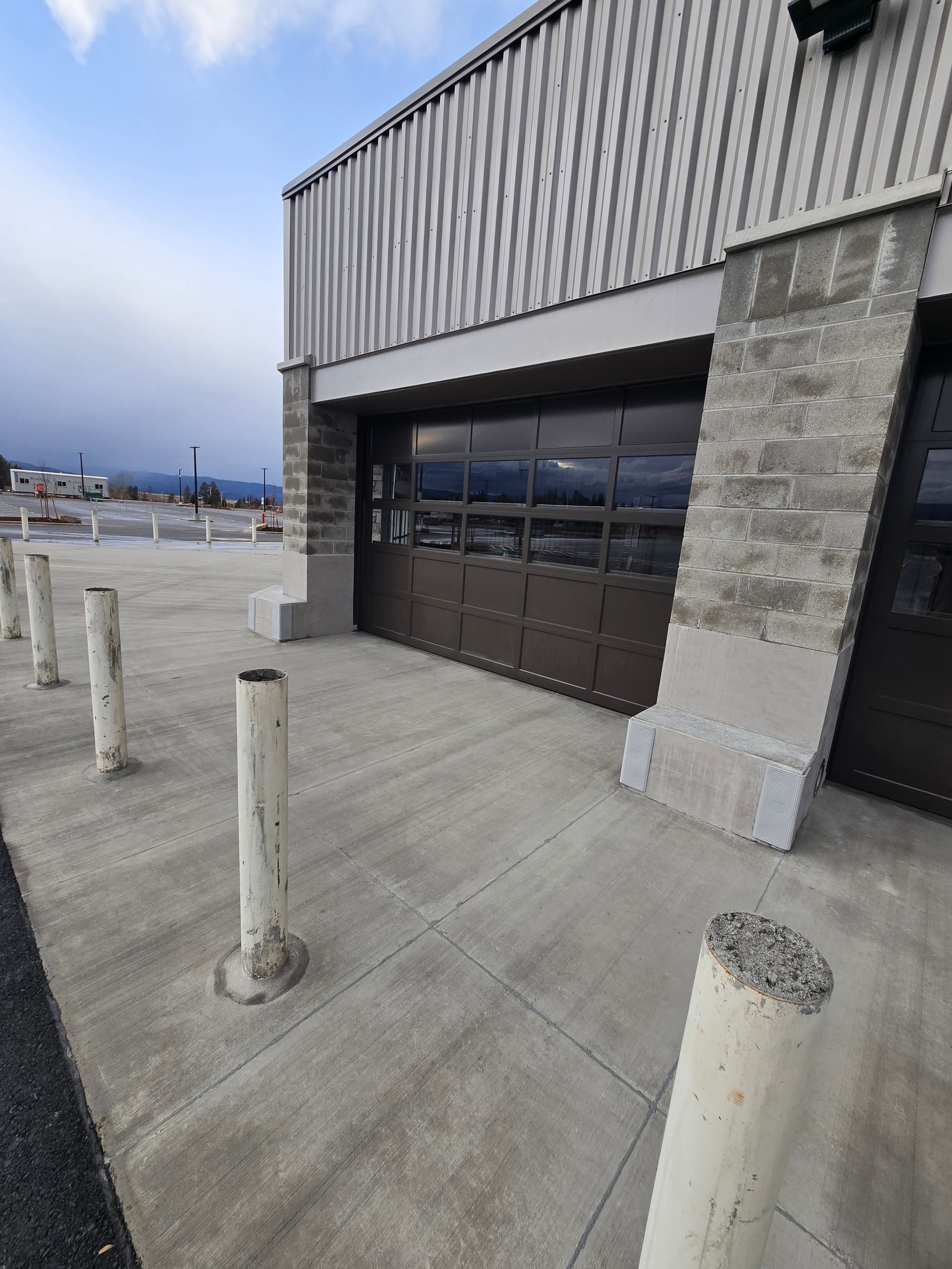 A brown sectional garage door on a tan building, with several white concrete bollards standing on the concrete pavement.