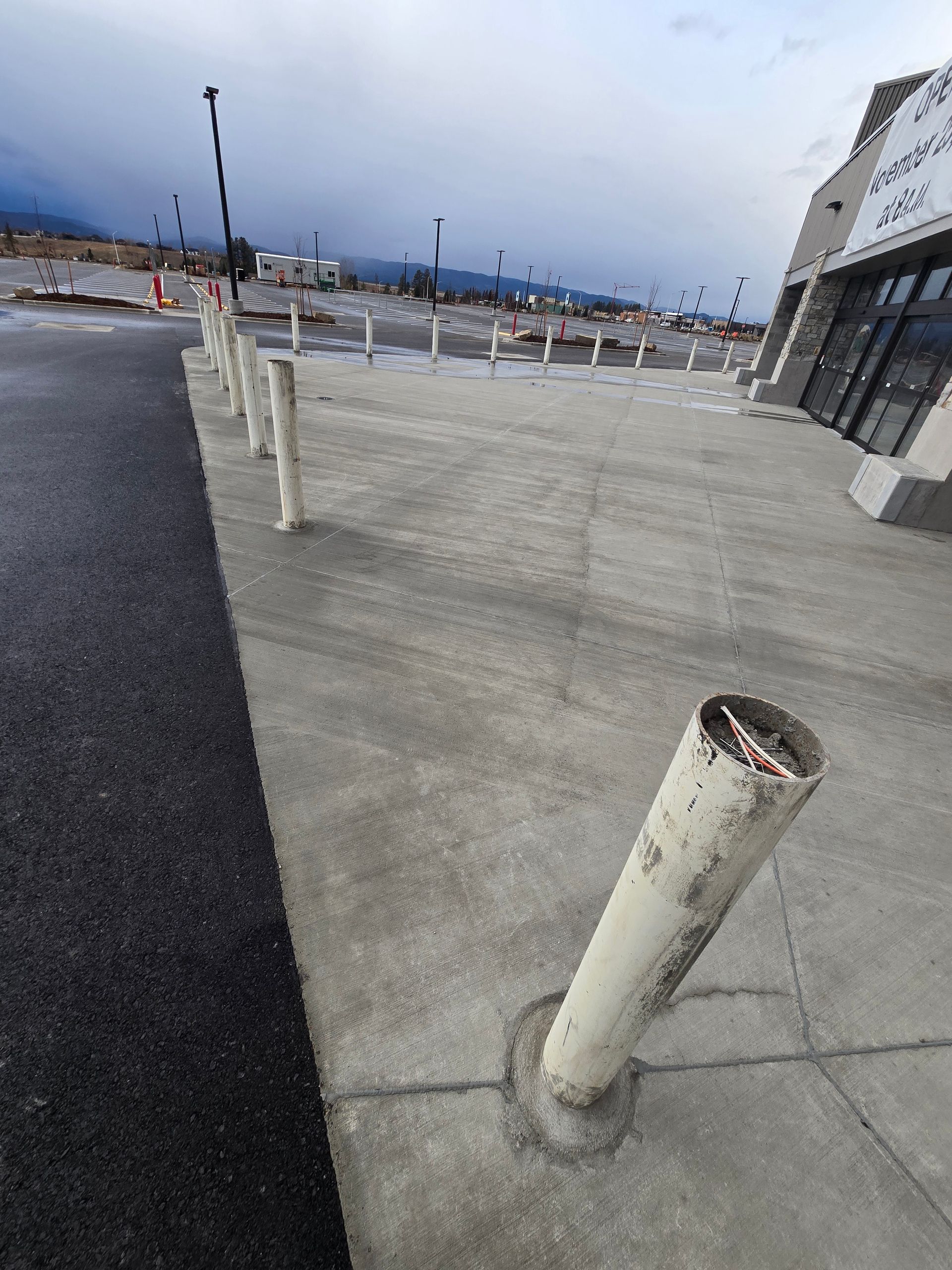 A row of white concrete bollards on a sidewalk next to an asphalt parking lot and a storefront.