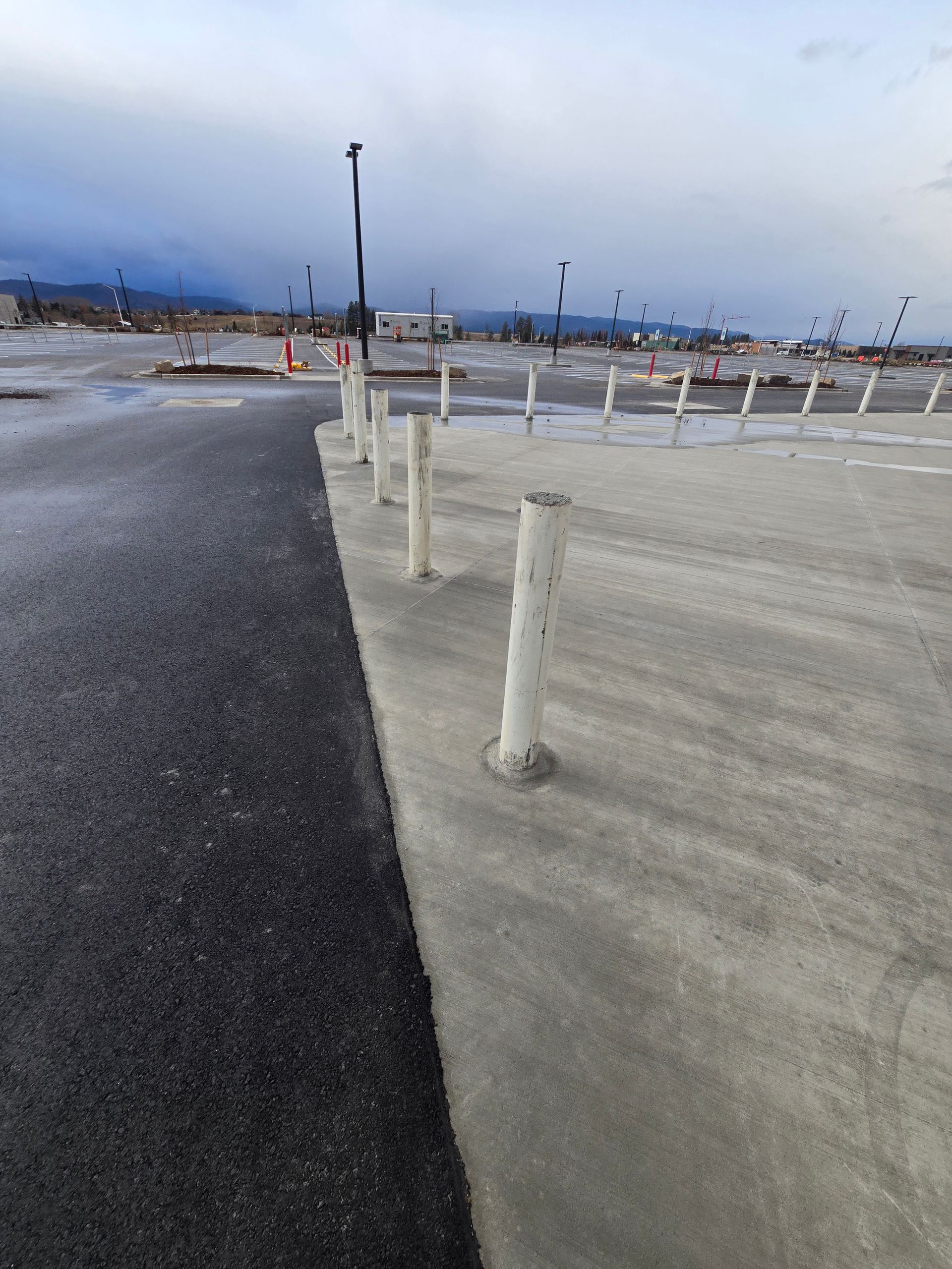 A row of white bollards separates an asphalt road from a concrete parking area under an overcast sky.
