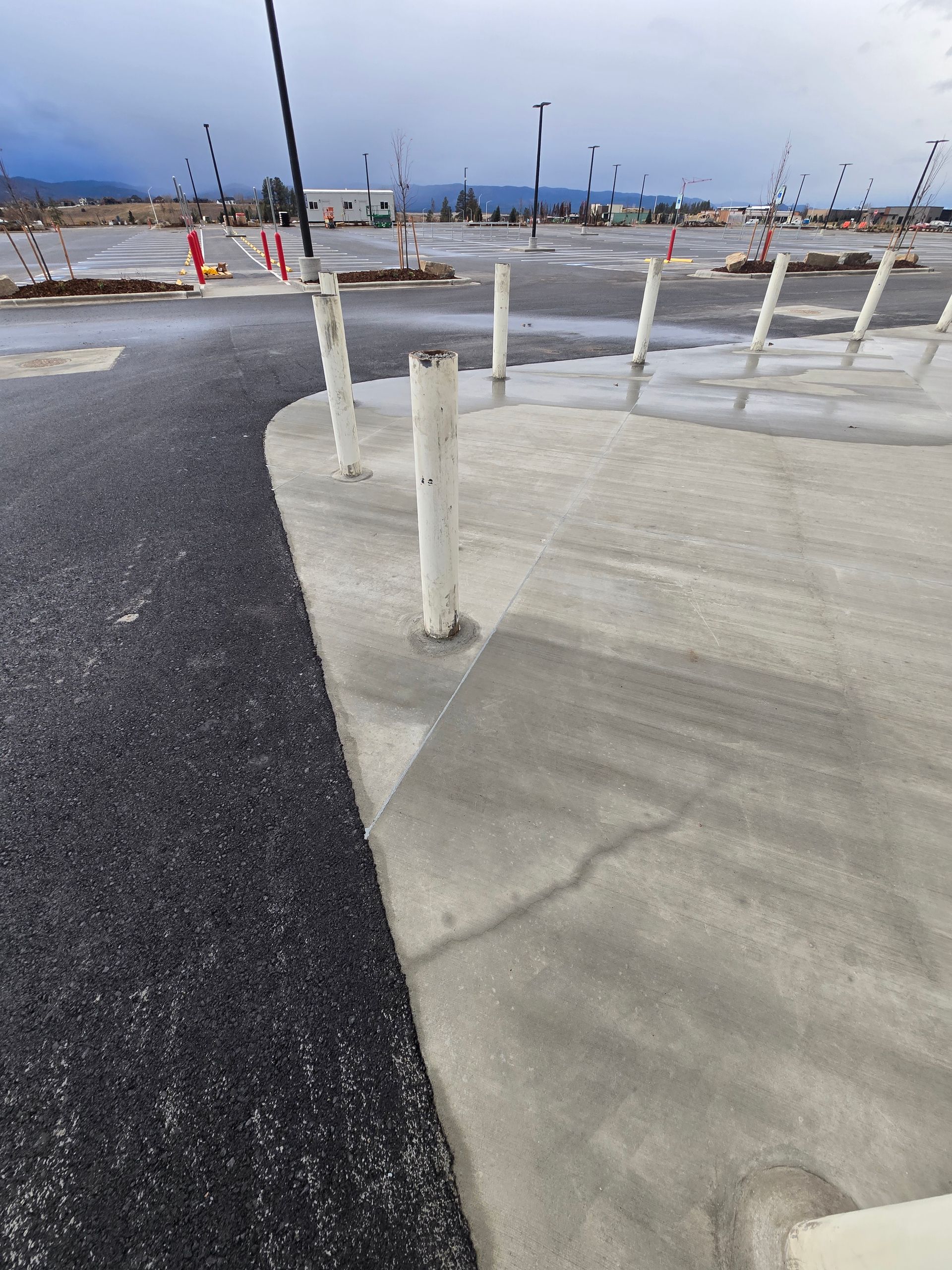 A row of white bollards separates an asphalt parking lot from a concrete walkway, with a crack visible in the concrete.