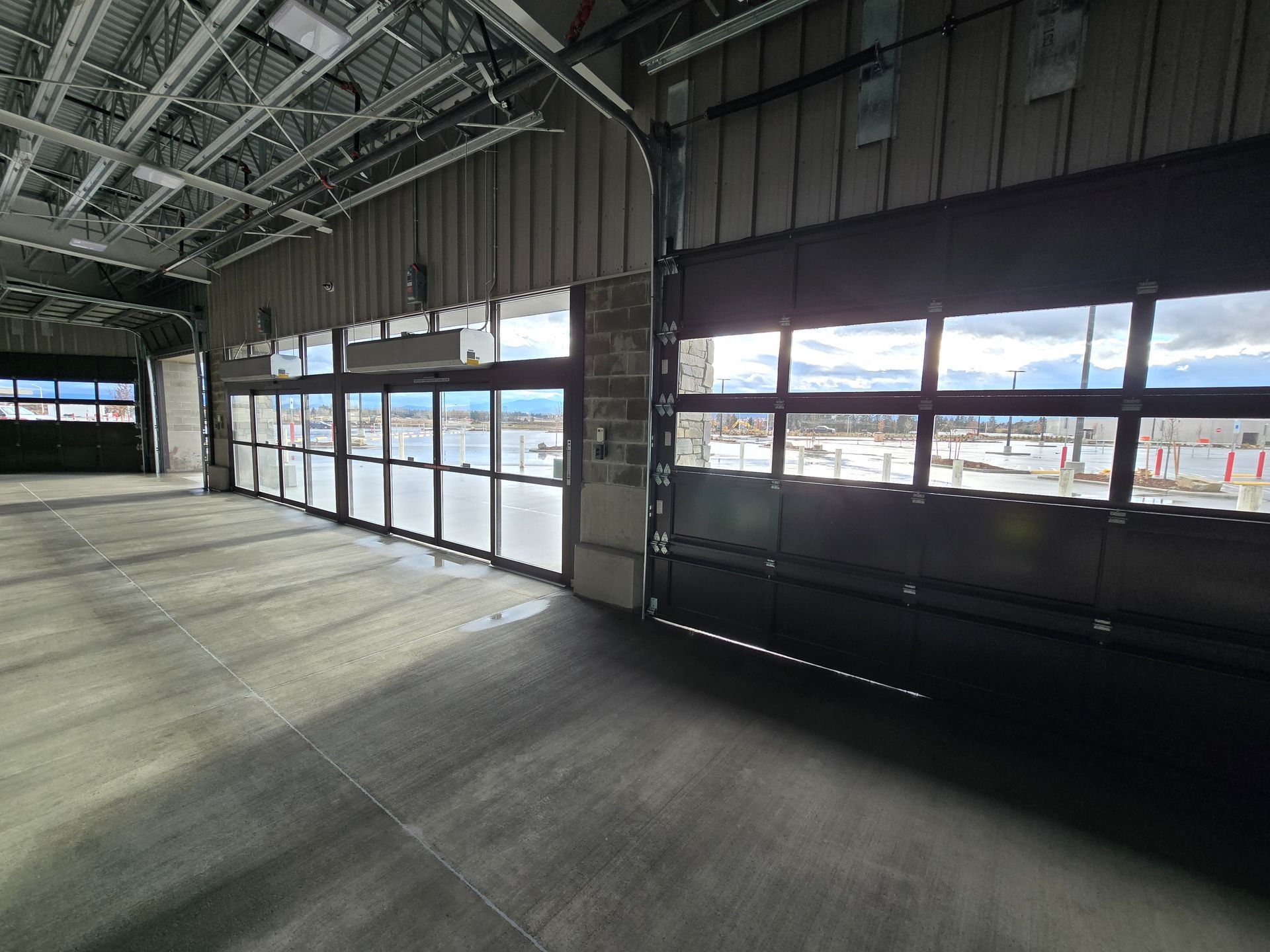 An empty industrial space with a concrete floor, high ceilings, and large glass garage doors looking out to a field.