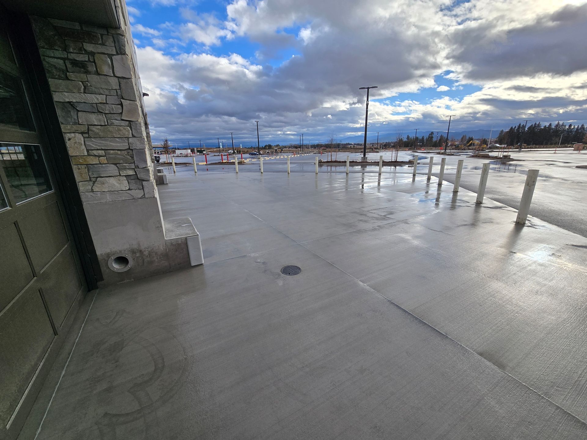 A wide, wet concrete patio outside a stone building, leading toward an open, cloudy parking area with metal bollards.