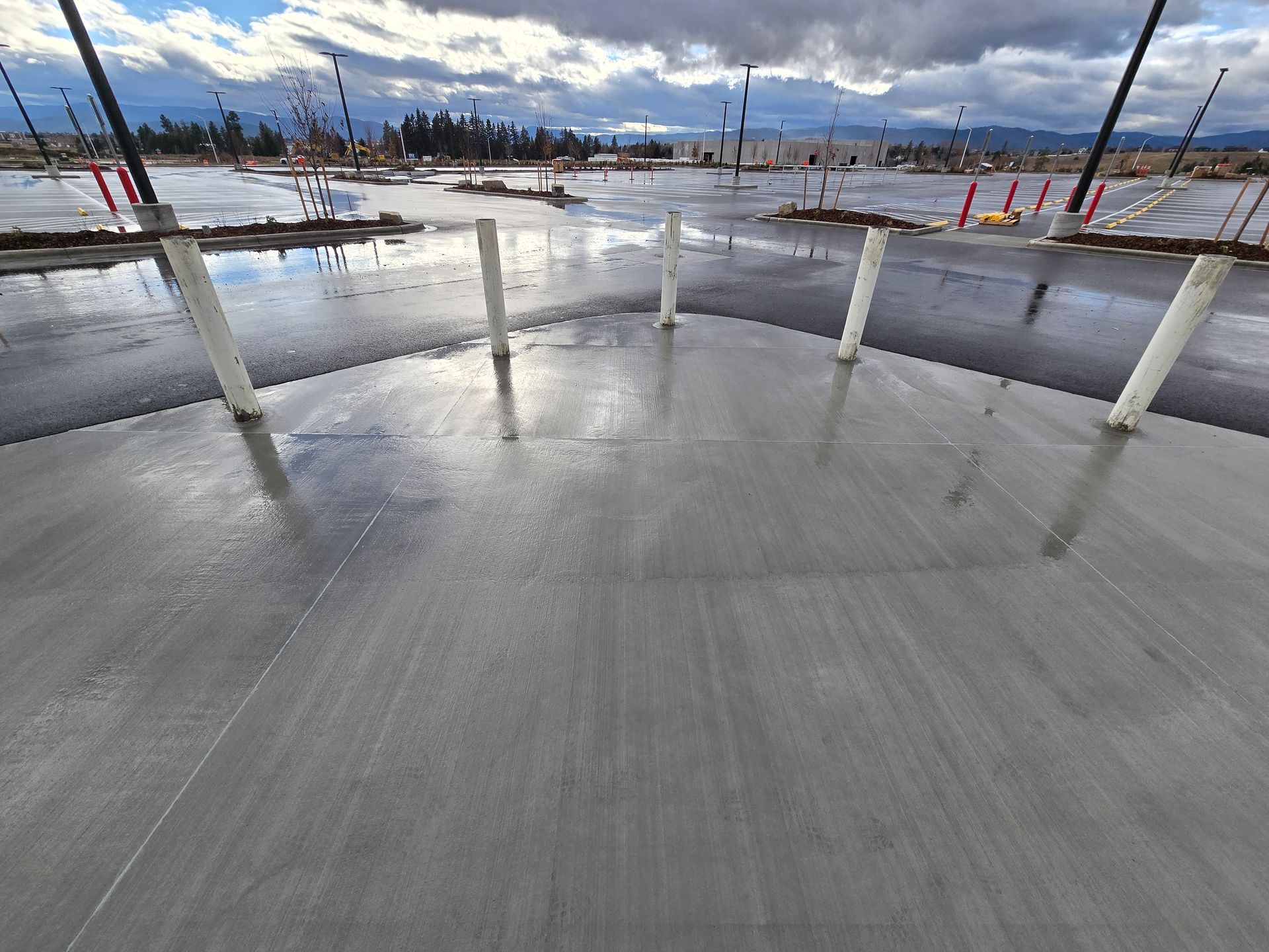 A row of white bollards stands on a newly poured concrete sidewalk near a wet, overcast parking lot.