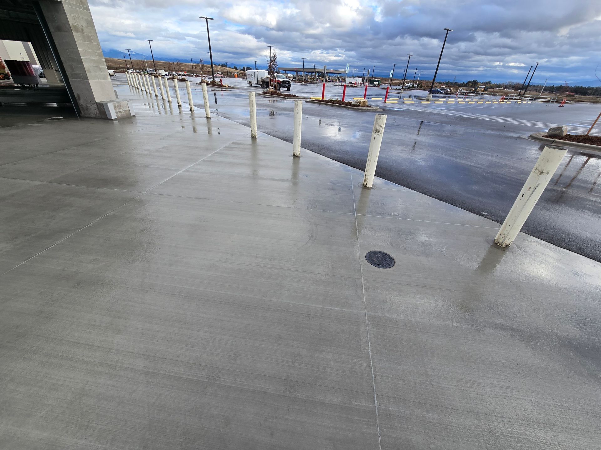 A row of white bollards lines a wet, gray concrete sidewalk next to a construction site under a cloudy sky.