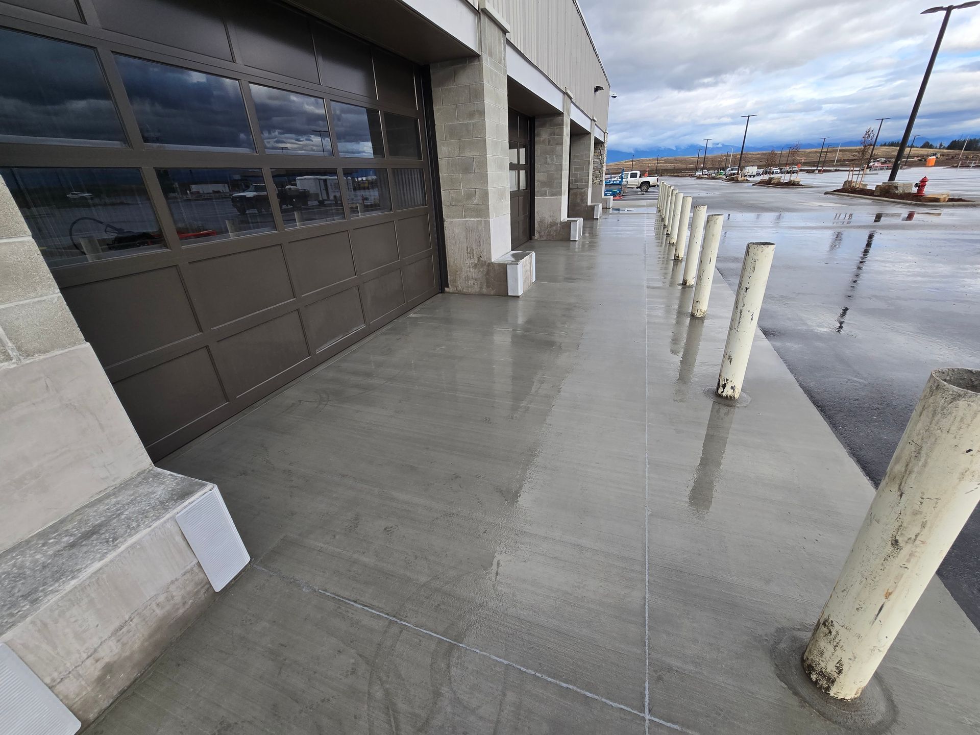 A concrete sidewalk alongside a building with a large garage door, lined with metal safety bollards on a rainy day.