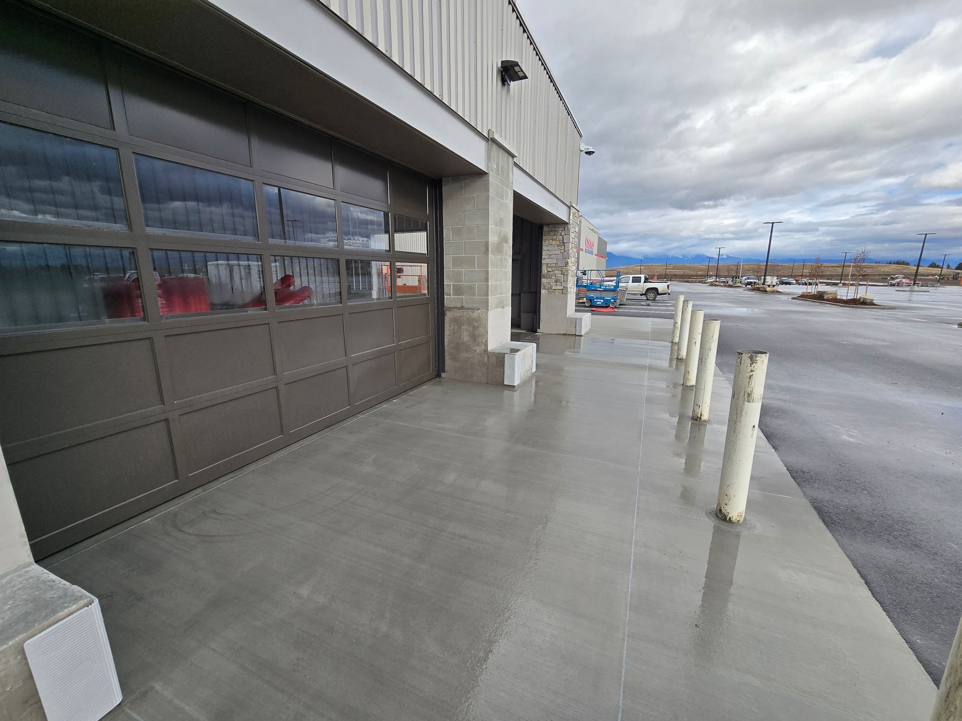 A modern building exterior featuring a large glass bay door, concrete walkway, and a row of safety bollards near a lot.