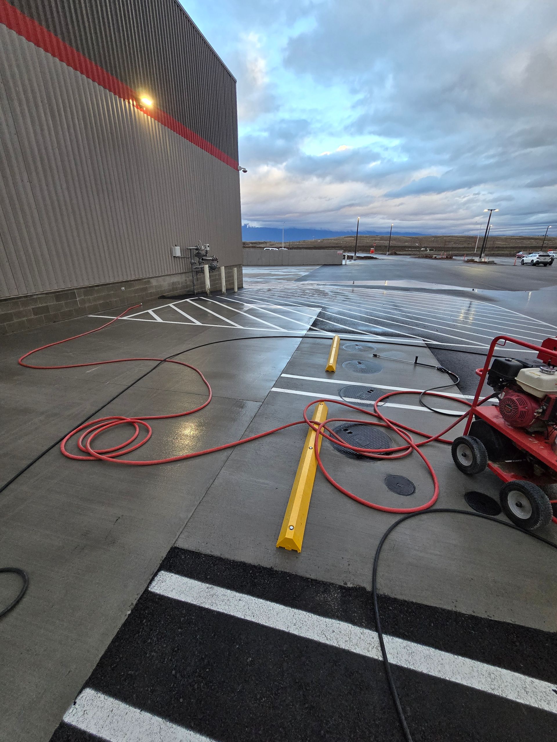 A pressure washer with red hoses cleans a parking lot’s painted crosswalk lines near a large industrial building.