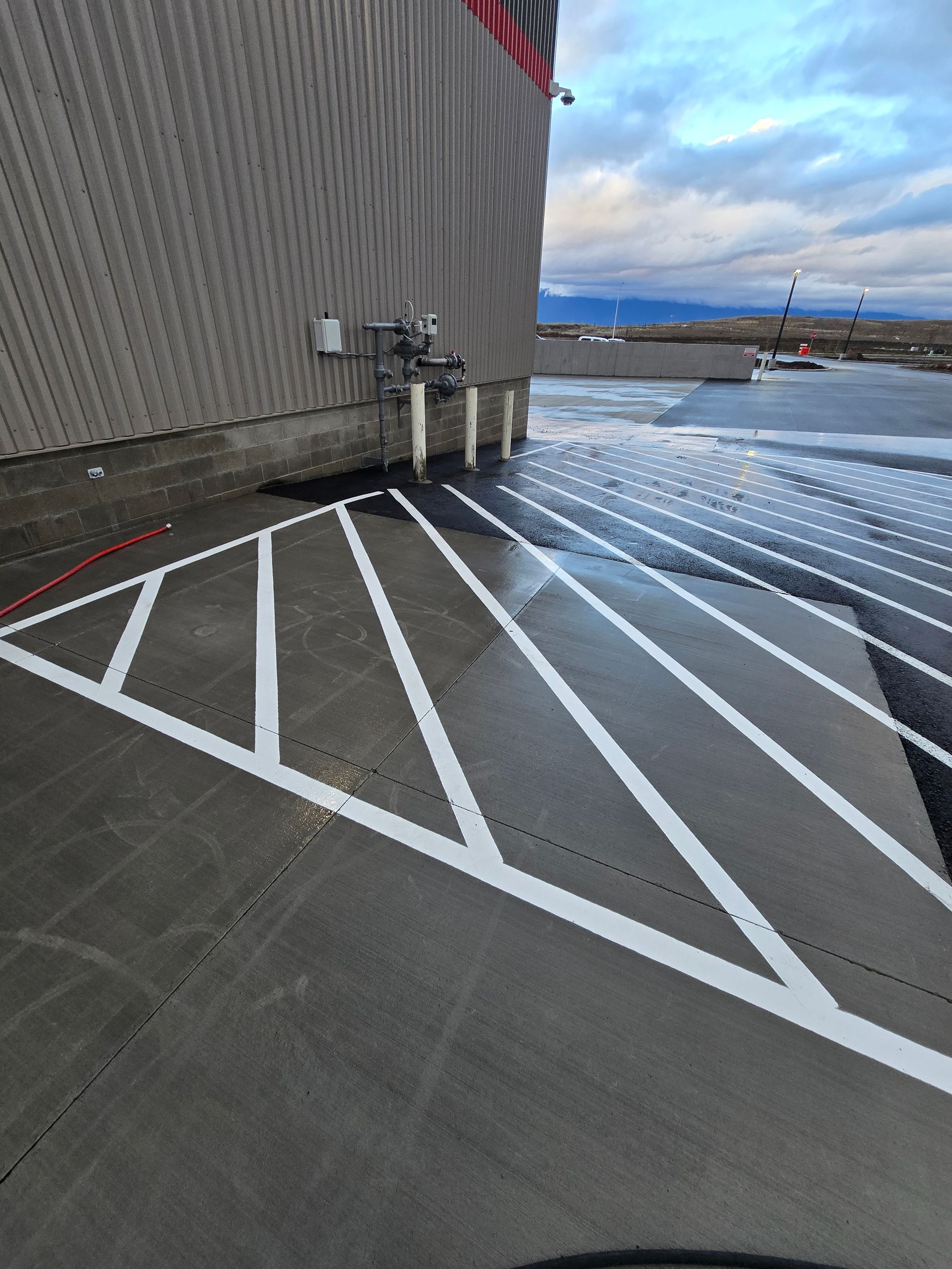 A paved area outside a building featuring white diagonal parking lot markings and a wet surface under a cloudy sky.