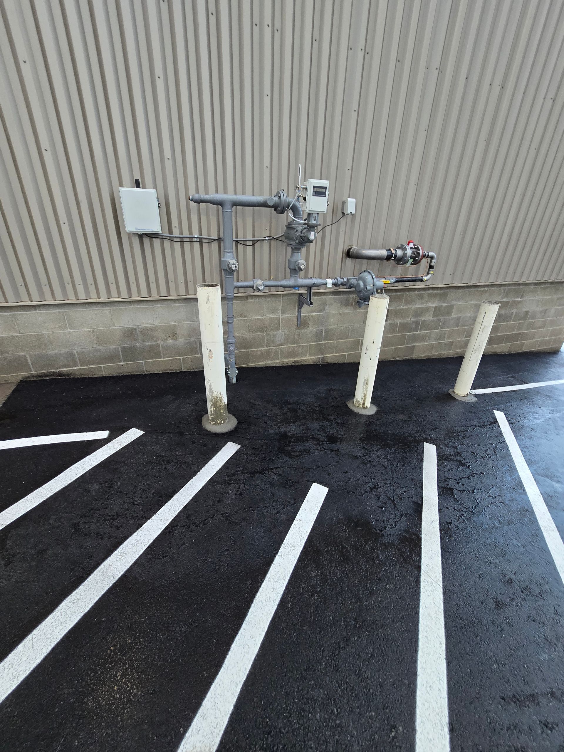 Gas meter assembly with protective bollards against a corrugated metal wall next to a paved parking lot.