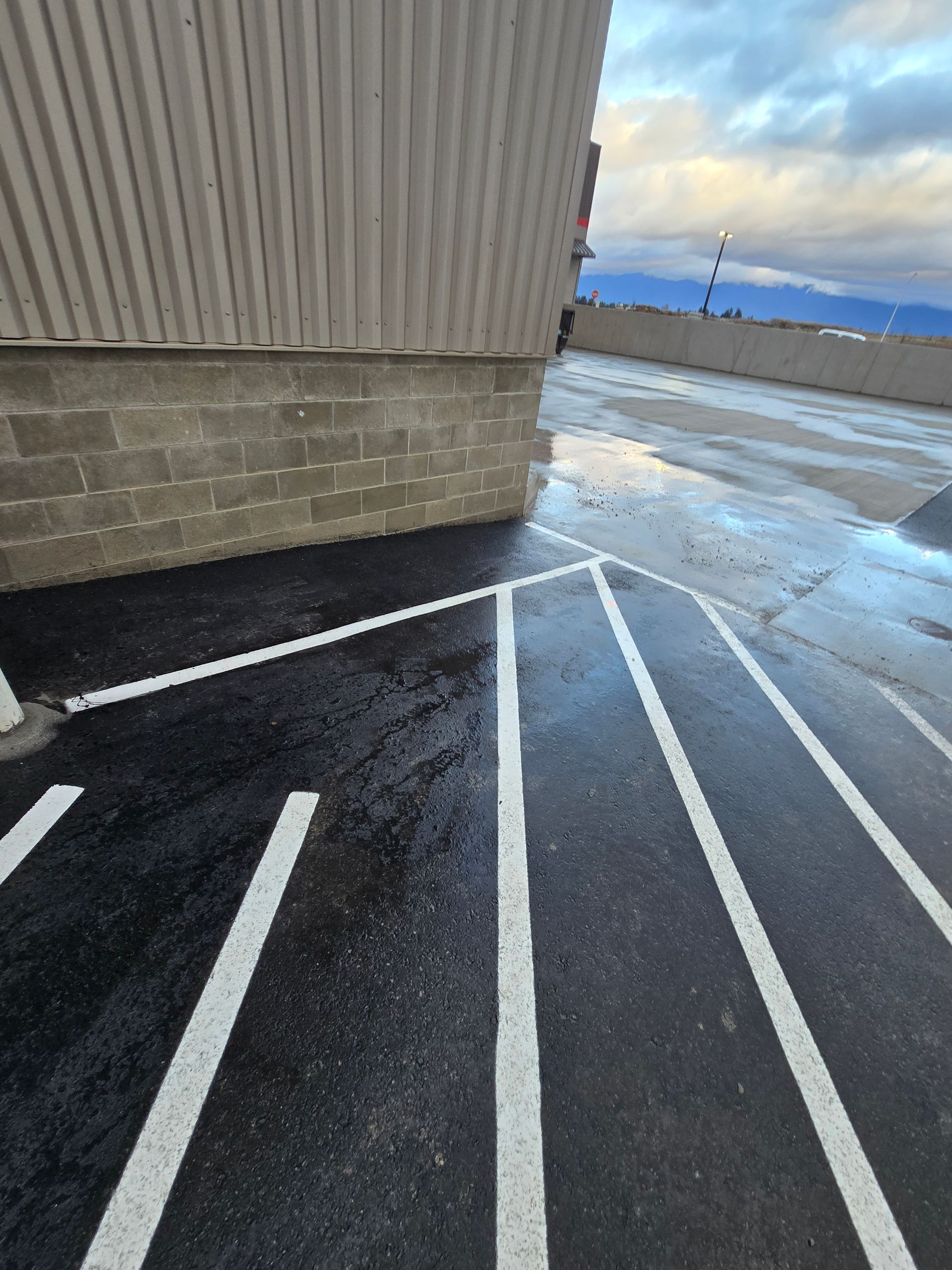 A corner of a building with light gray metal siding and a brick base next to a freshly painted parking lot with white lines.