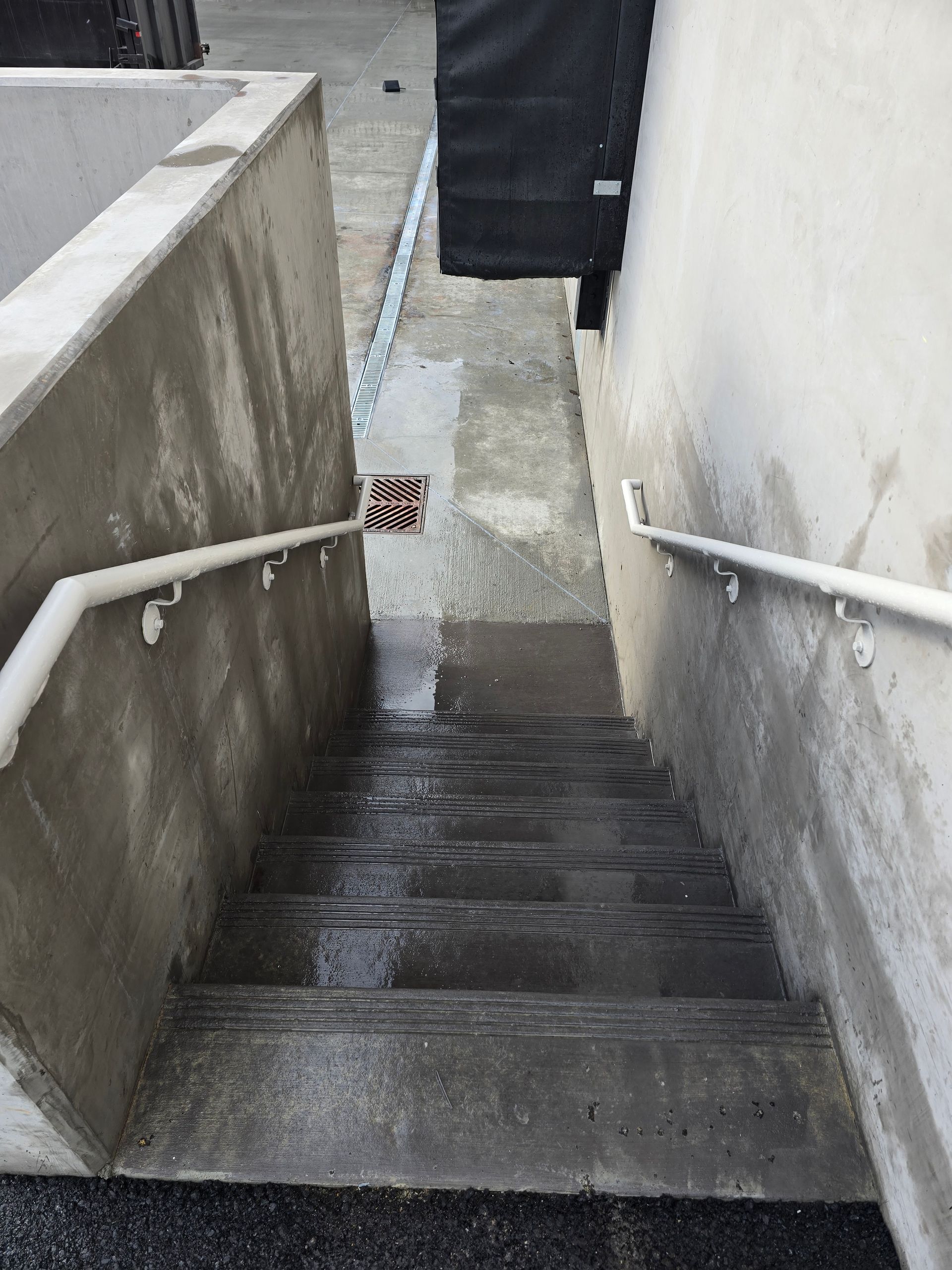 An outdoor concrete staircase descends between two white walls, featuring white handrails on both sides.