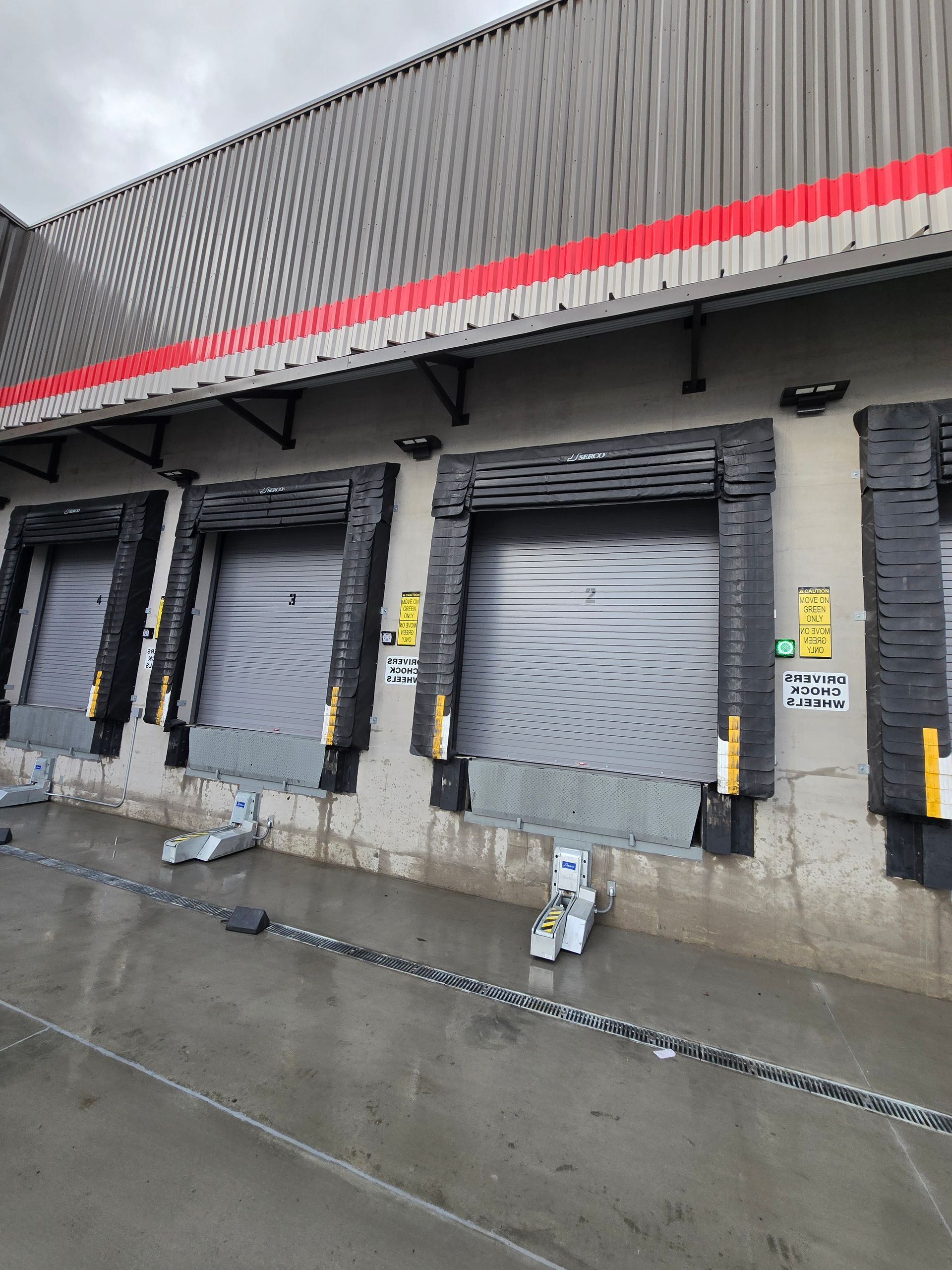 A row of four industrial loading dock bays with closed metal doors and black rubber bumpers on a concrete building.