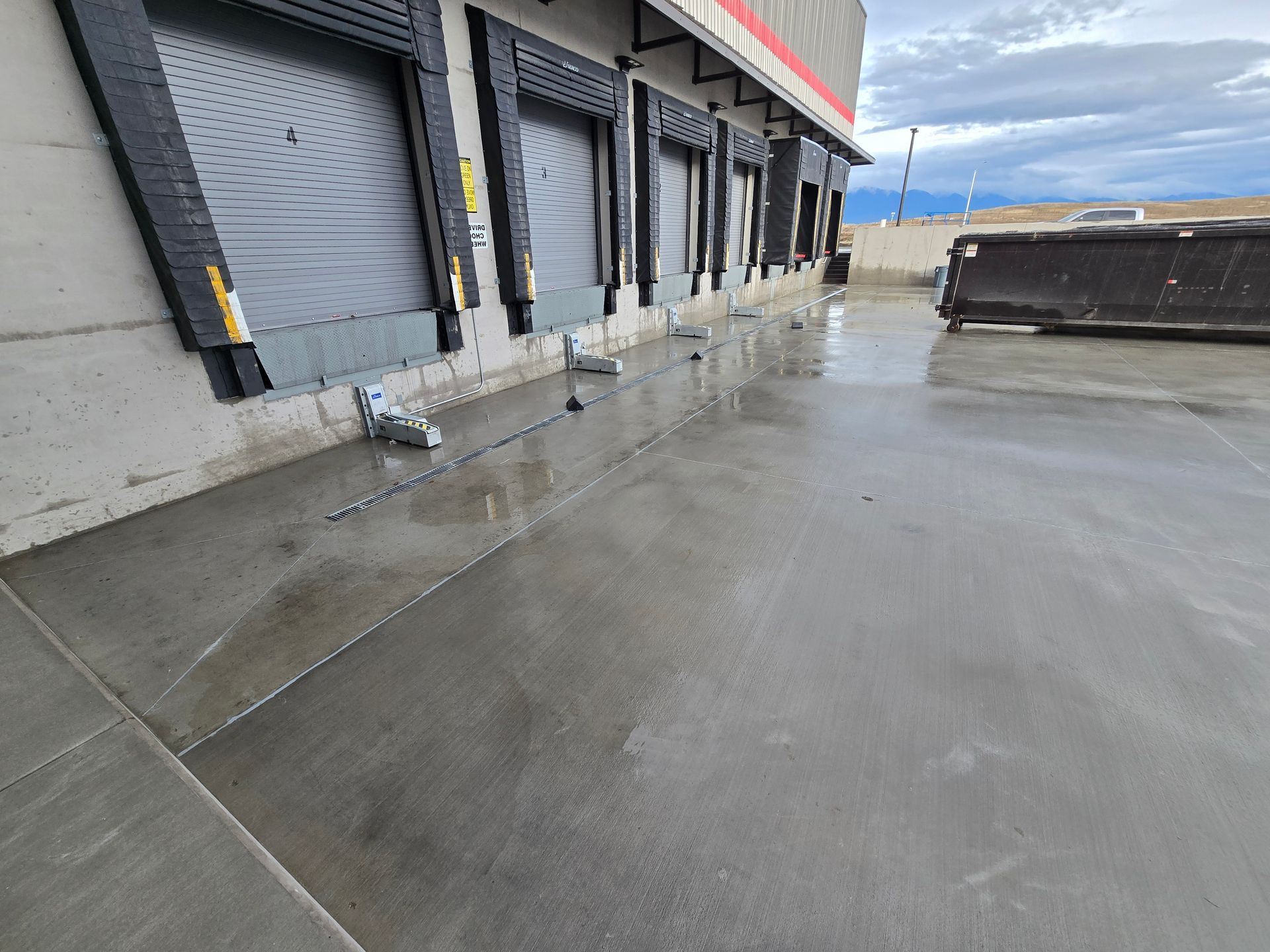 Loading dock area with multiple closed roll-up doors and a large dumpster on a concrete lot under a cloudy sky.