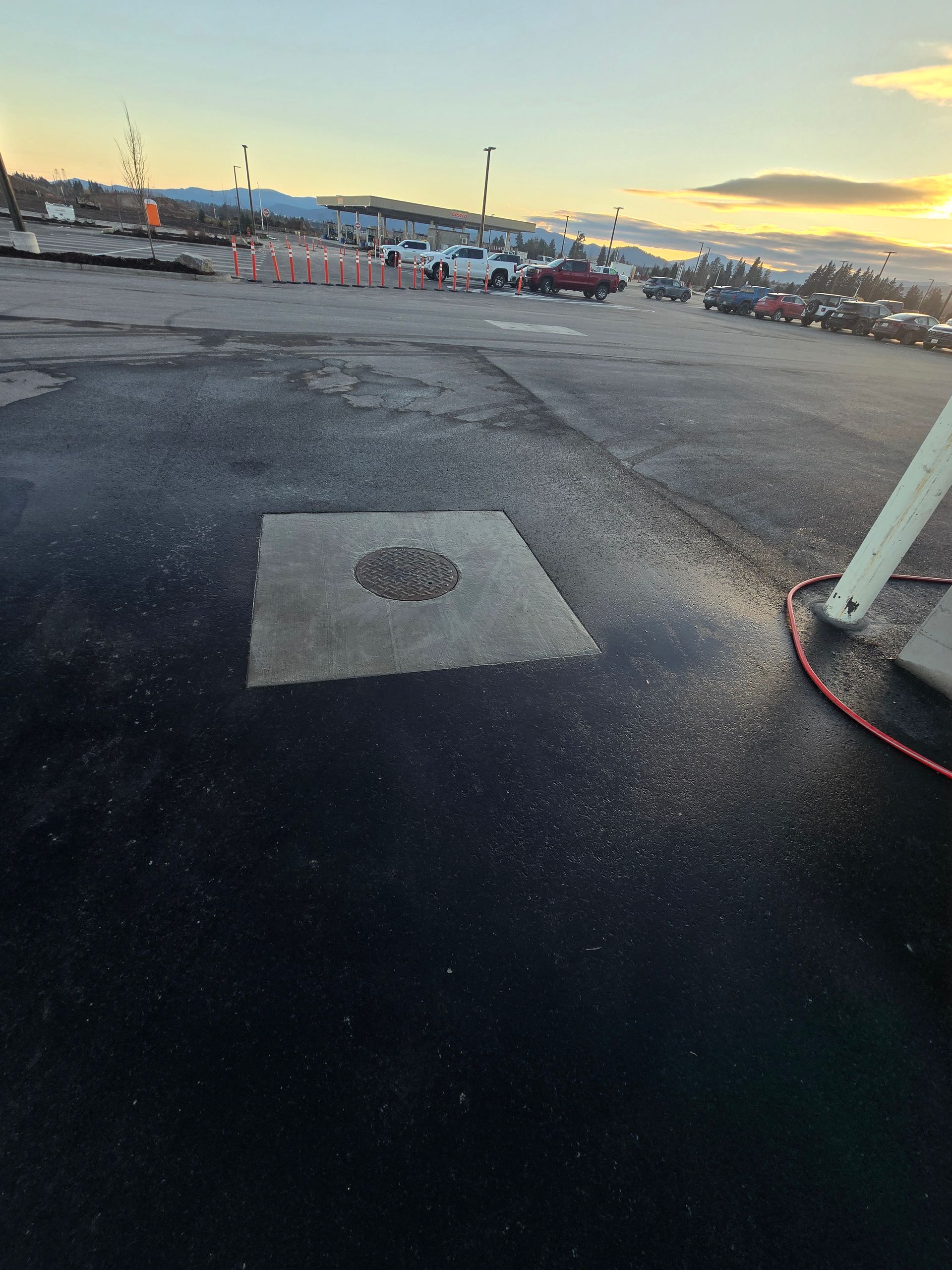 A square concrete patch with a circular drain cover sits in a dark asphalt parking lot at sunset near a gas station.