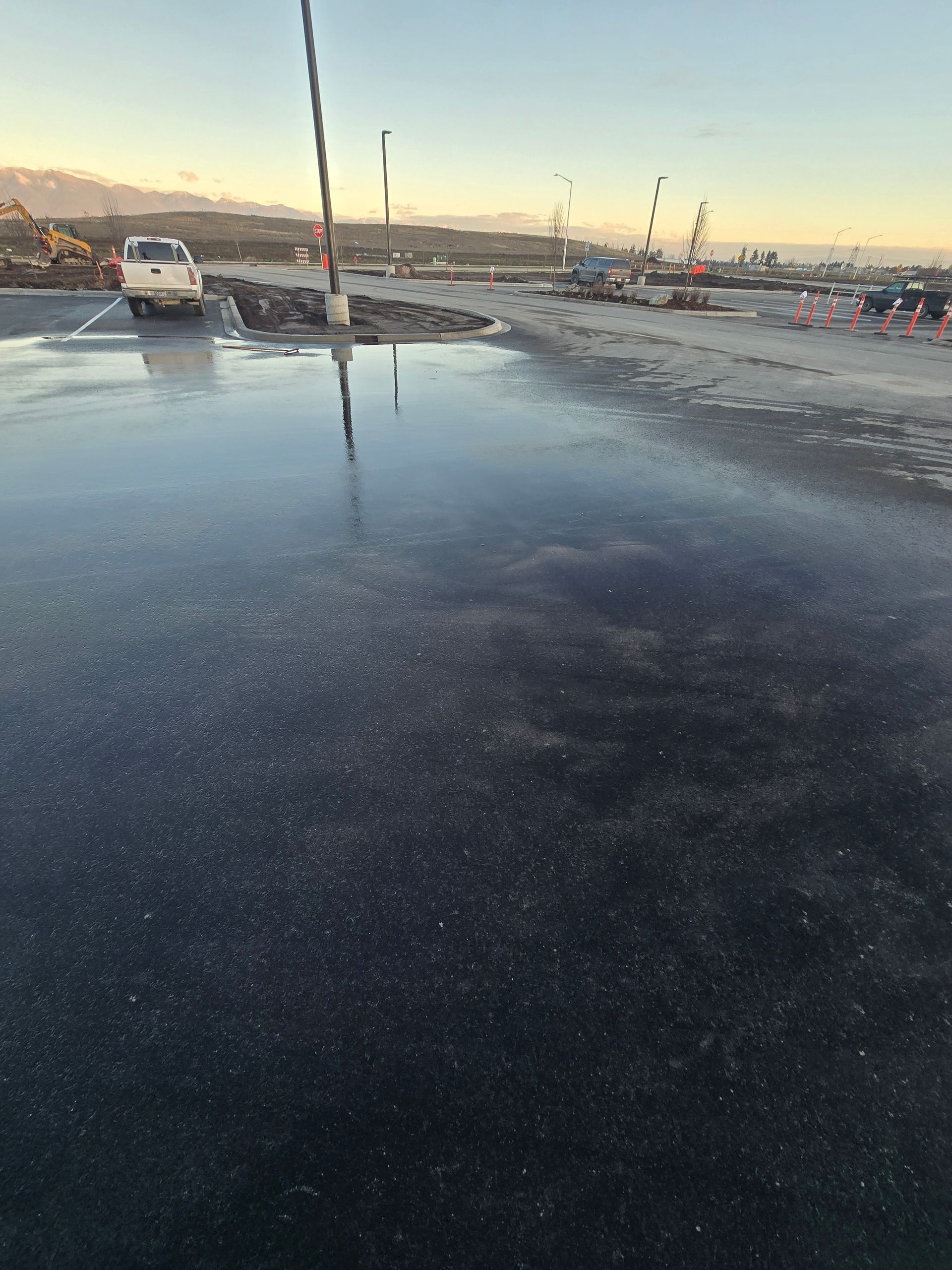 A large puddle reflecting a light-colored truck and a thin pole on a paved surface at sunset.