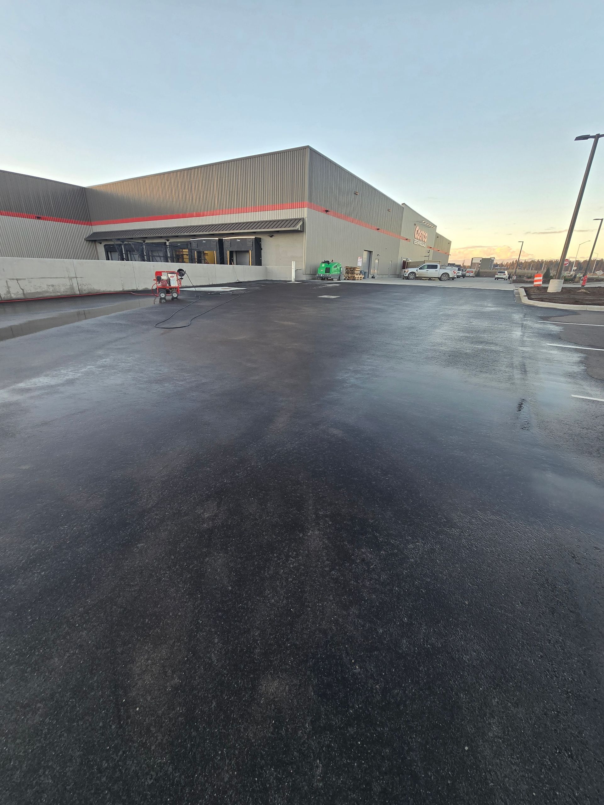A large, modern warehouse-style building with patterned walls overlooks a wet, empty asphalt parking lot at sunset.