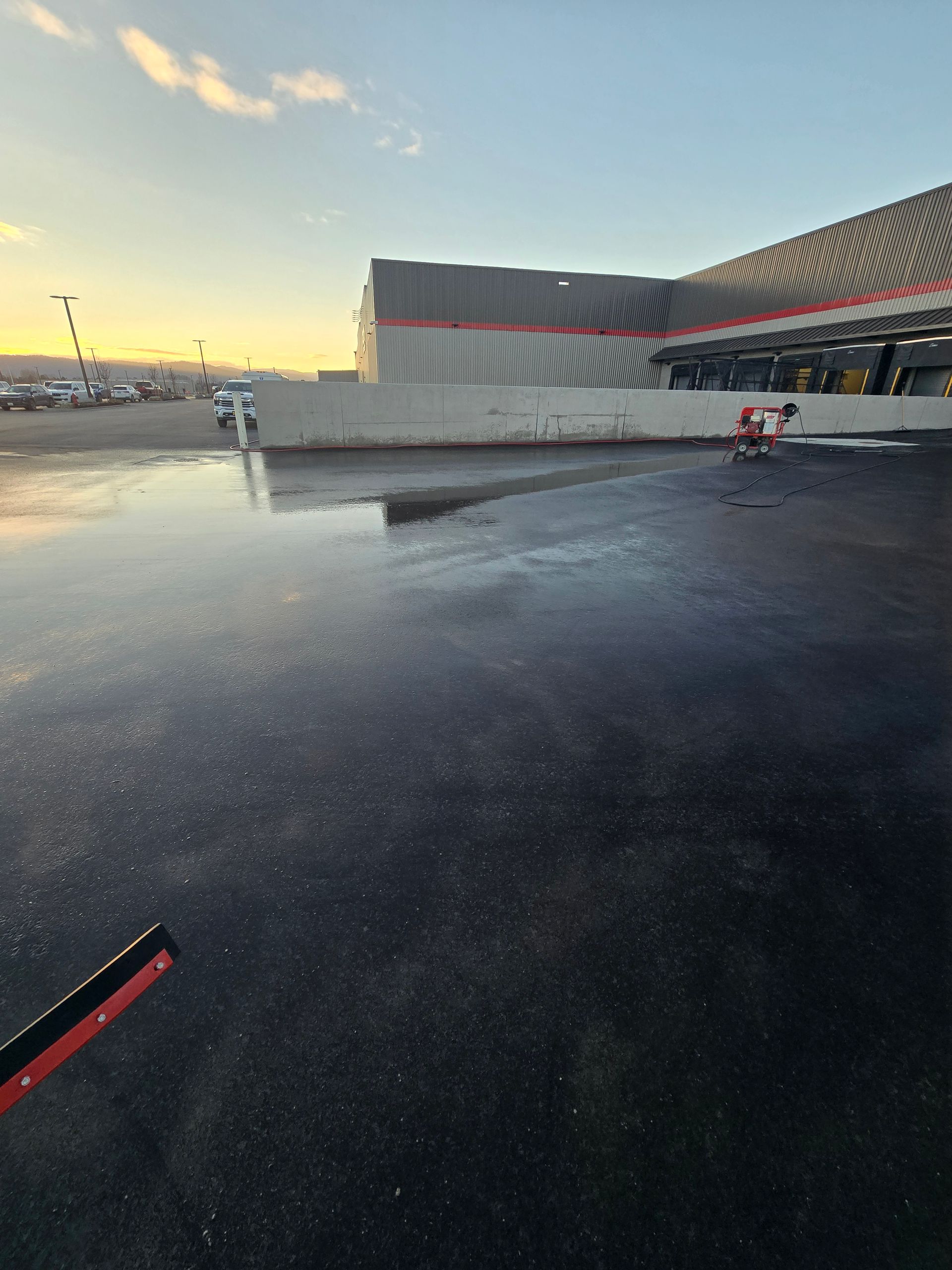 A sunset view of a wet, reflective parking lot next to a large warehouse building with a solitary shopping cart nearby.