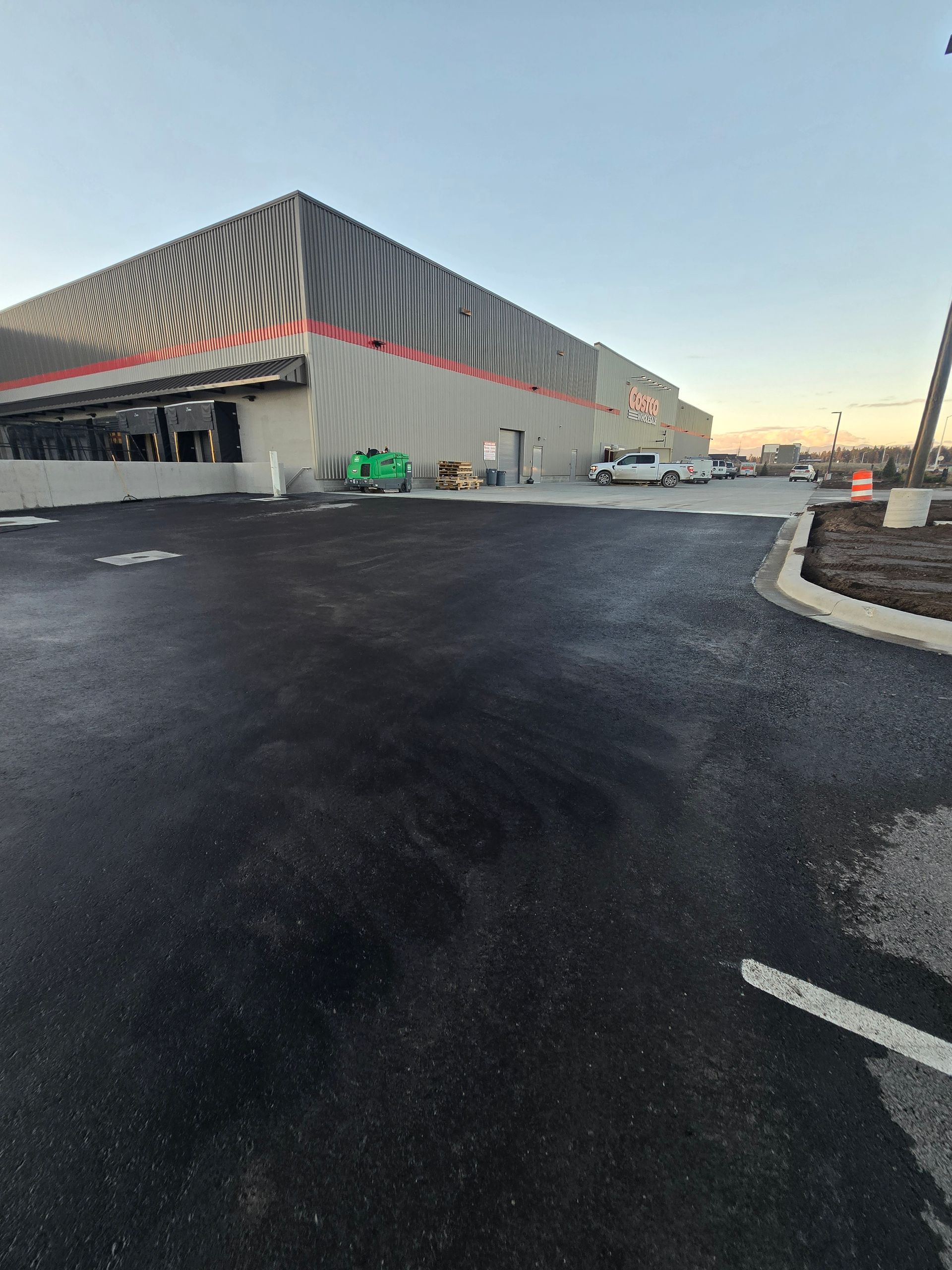 A modern industrial warehouse with grey walls and red striping, featuring a newly paved asphalt lot in the foreground.