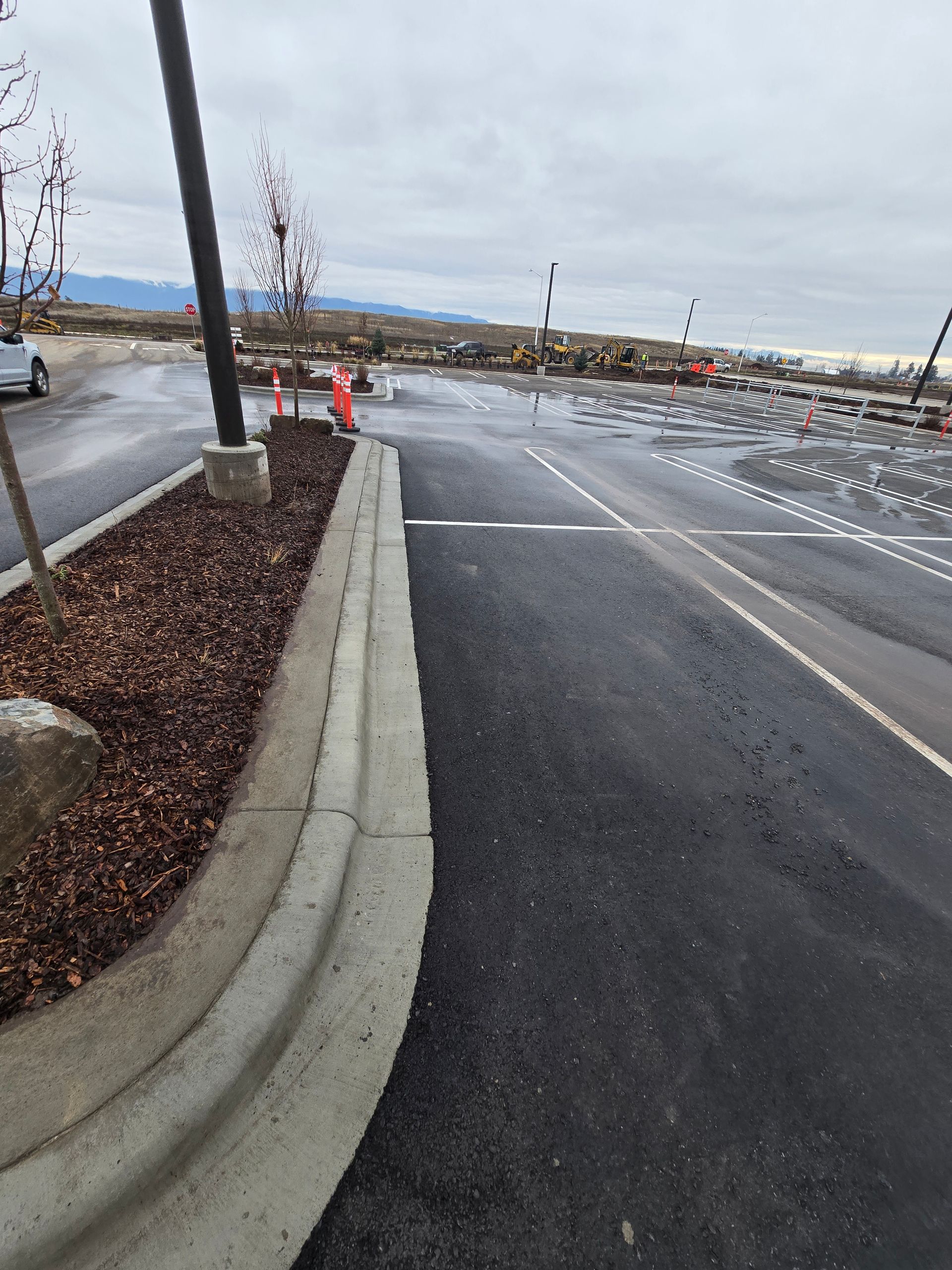 A paved parking lot with a raised concrete curb, mulch island, and parking spaces under a cloudy sky.