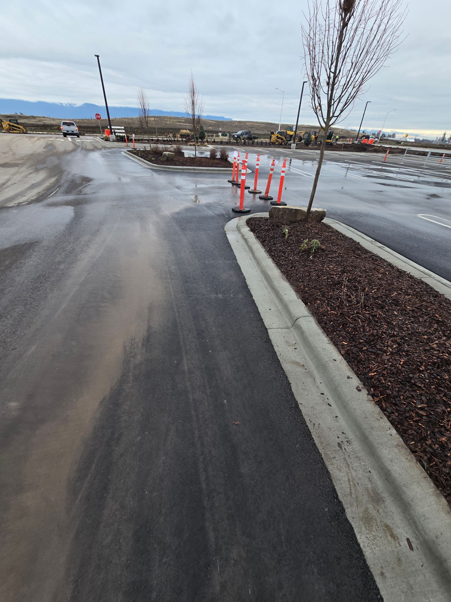 A paved parking lot with a landscaped curb and construction cones, showing muddy tire tracks and distant heavy machinery.