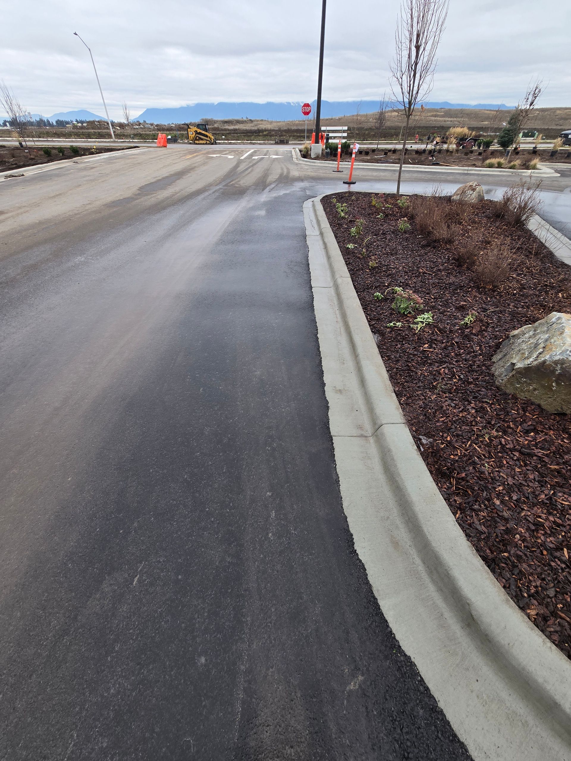 A paved road curves alongside a landscaped median with mulch and a rock, leading toward an open area under a cloudy sky.
