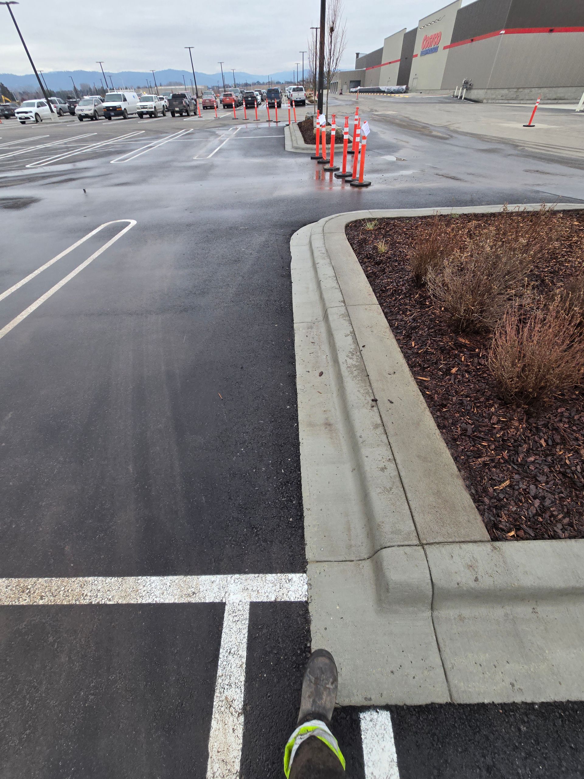 A person's foot in a work boot stands on asphalt next to a concrete curb in a parking lot with orange traffic cones ahead.