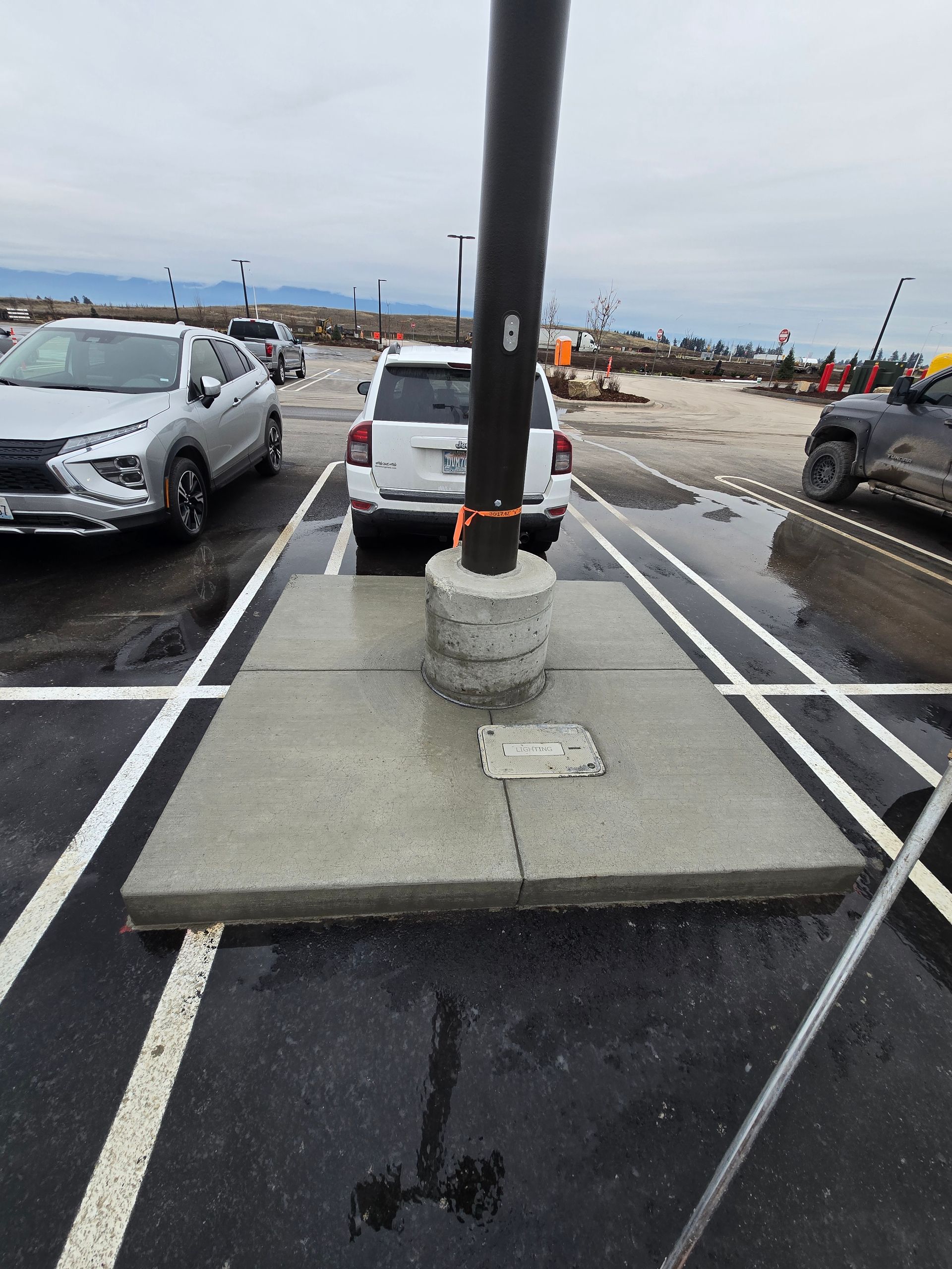 A dark light pole rises from a rectangular concrete island in a parking lot between two parked cars on a cloudy day.
