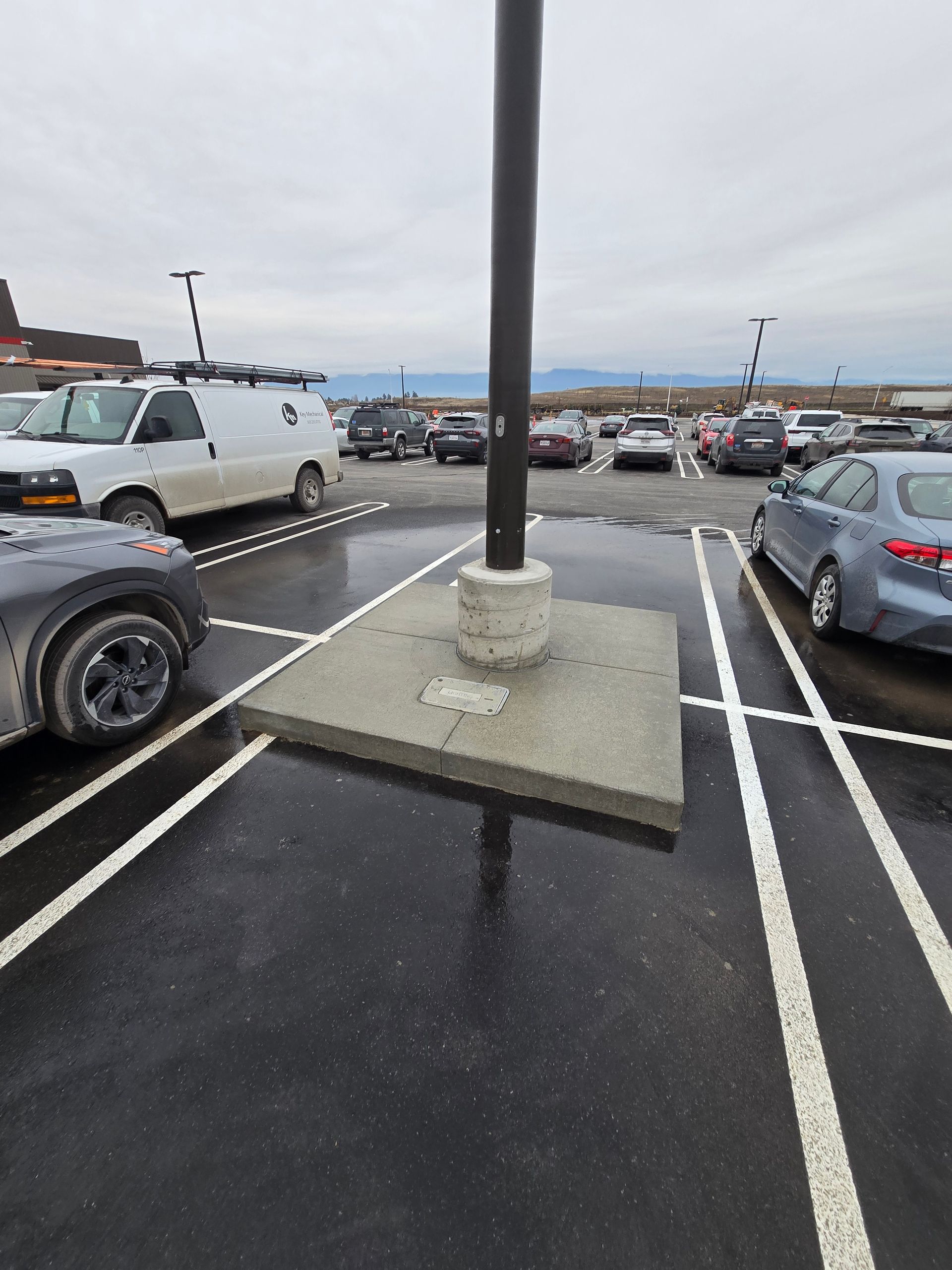 A concrete island with a light pole sits in a parking lot between two rows of parked vehicles on an overcast day.