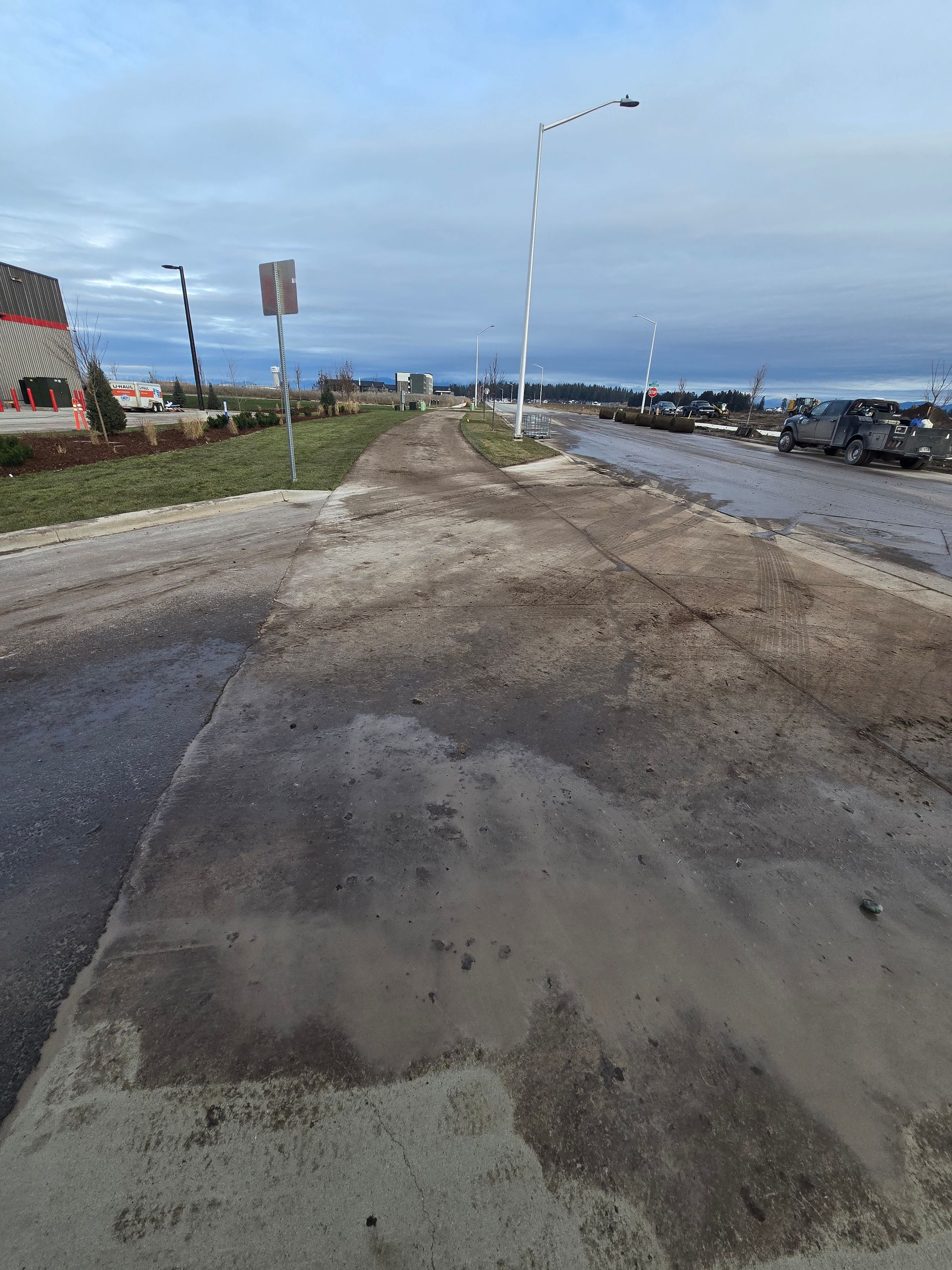 A muddy, unpaved construction site road stretches into the distance under a cloudy sky, with streetlights to the side.