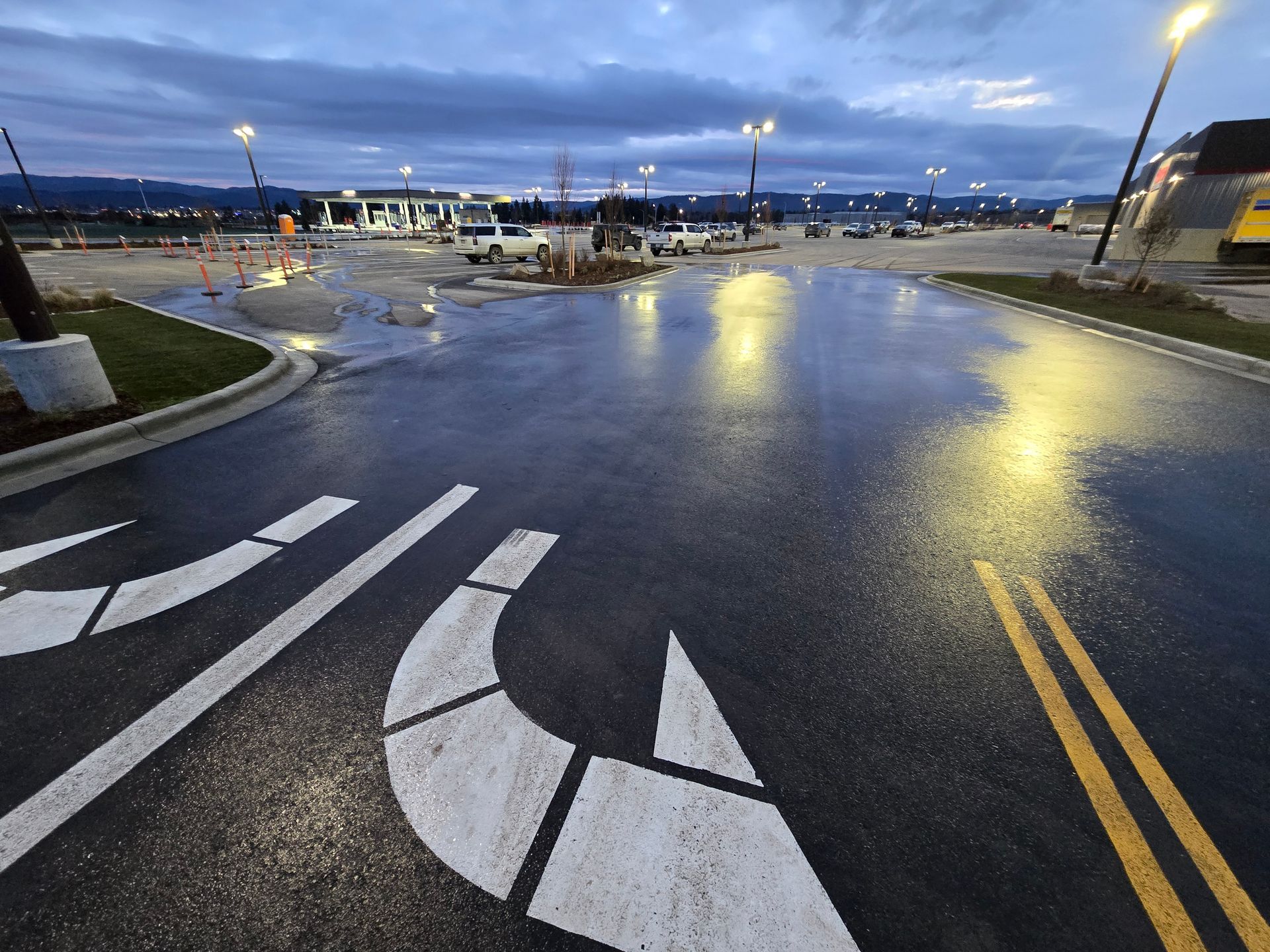A wet, dark parking lot at dusk with painted white lane markings and yellow traffic lines under outdoor streetlights.