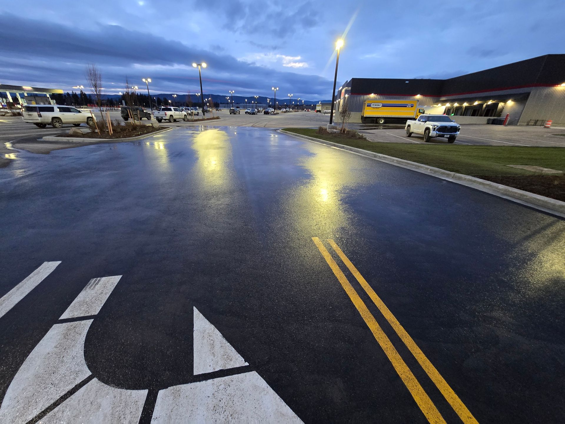 Wet pavement in a parking lot at twilight, with yellow lane markings, a white directional arrow, and parked cars.