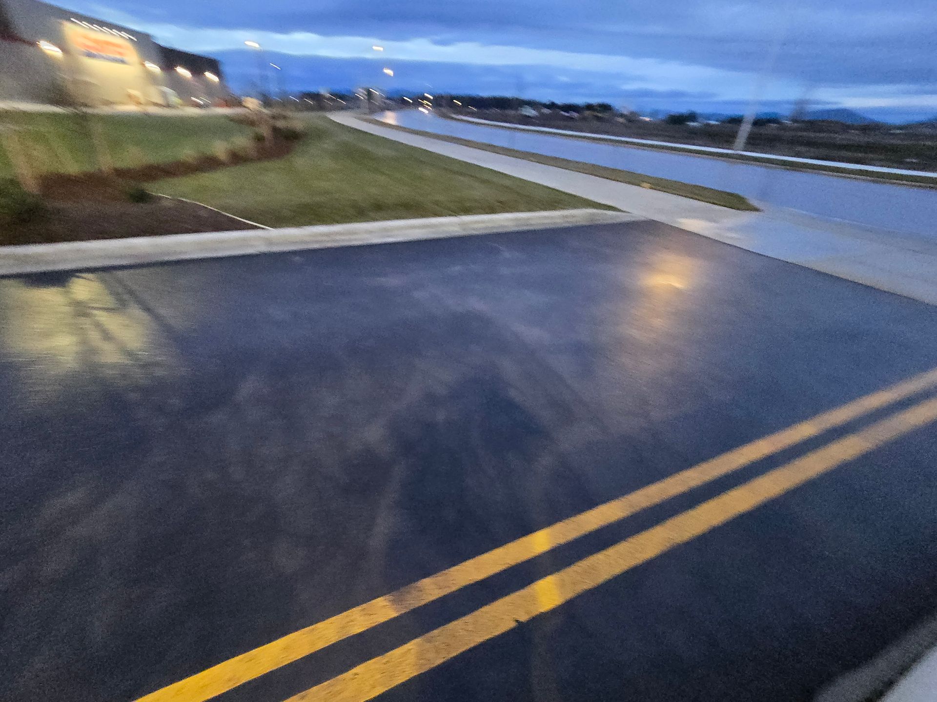 A parking lot or driveway with double yellow painted lines at dusk, with a building and landscape in the background.
