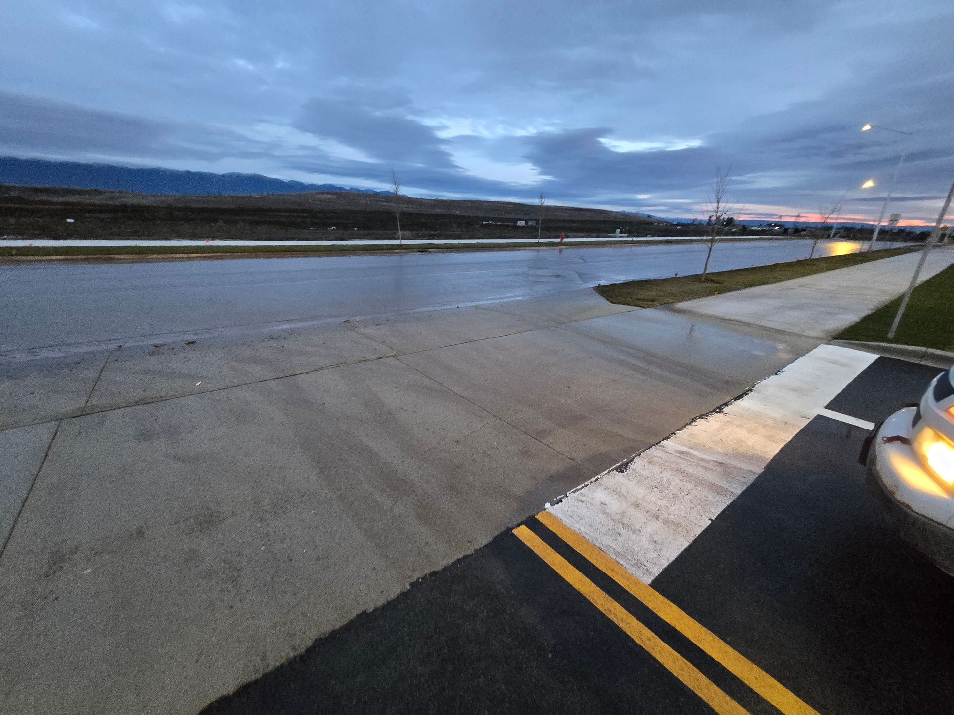 A wet paved road stretching into the distance at dusk, viewed from the driver's side of a parked vehicle.