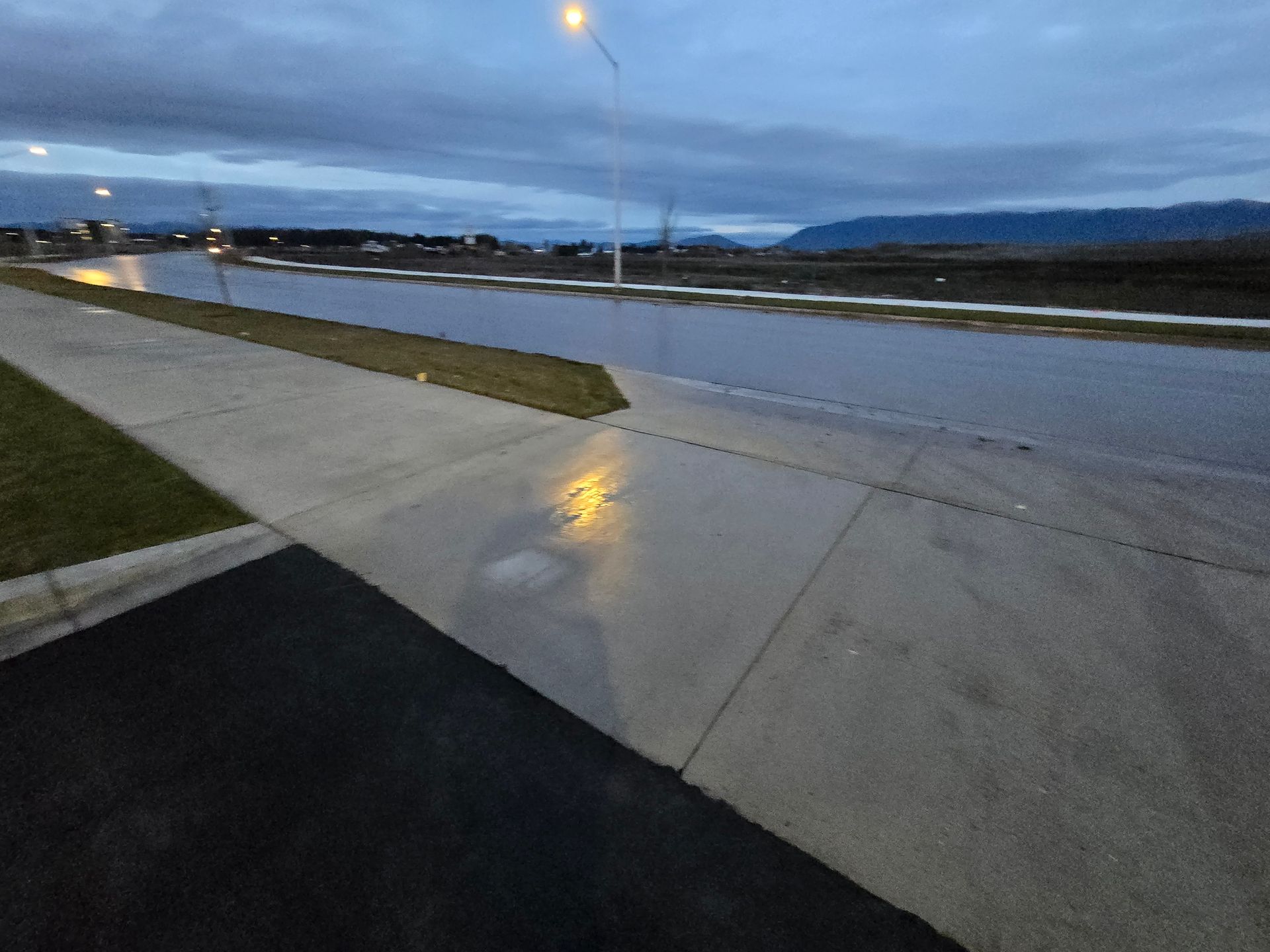 A street scene at dusk with a flooded road, a concrete sidewalk, and a streetlamp reflection on the wet pavement.