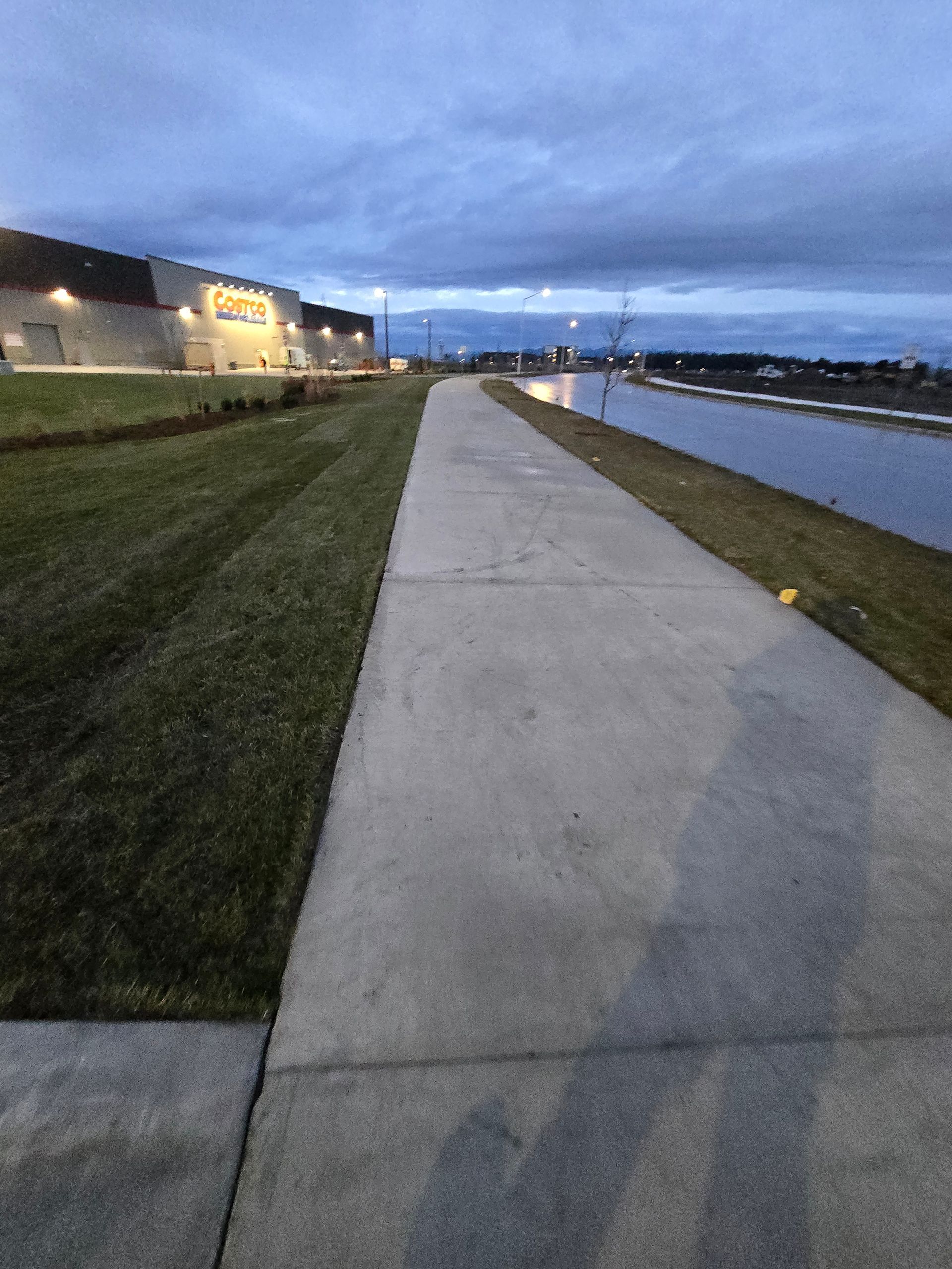 A paved path runs between a grassy area by a large industrial building and a body of water under a twilight sky.