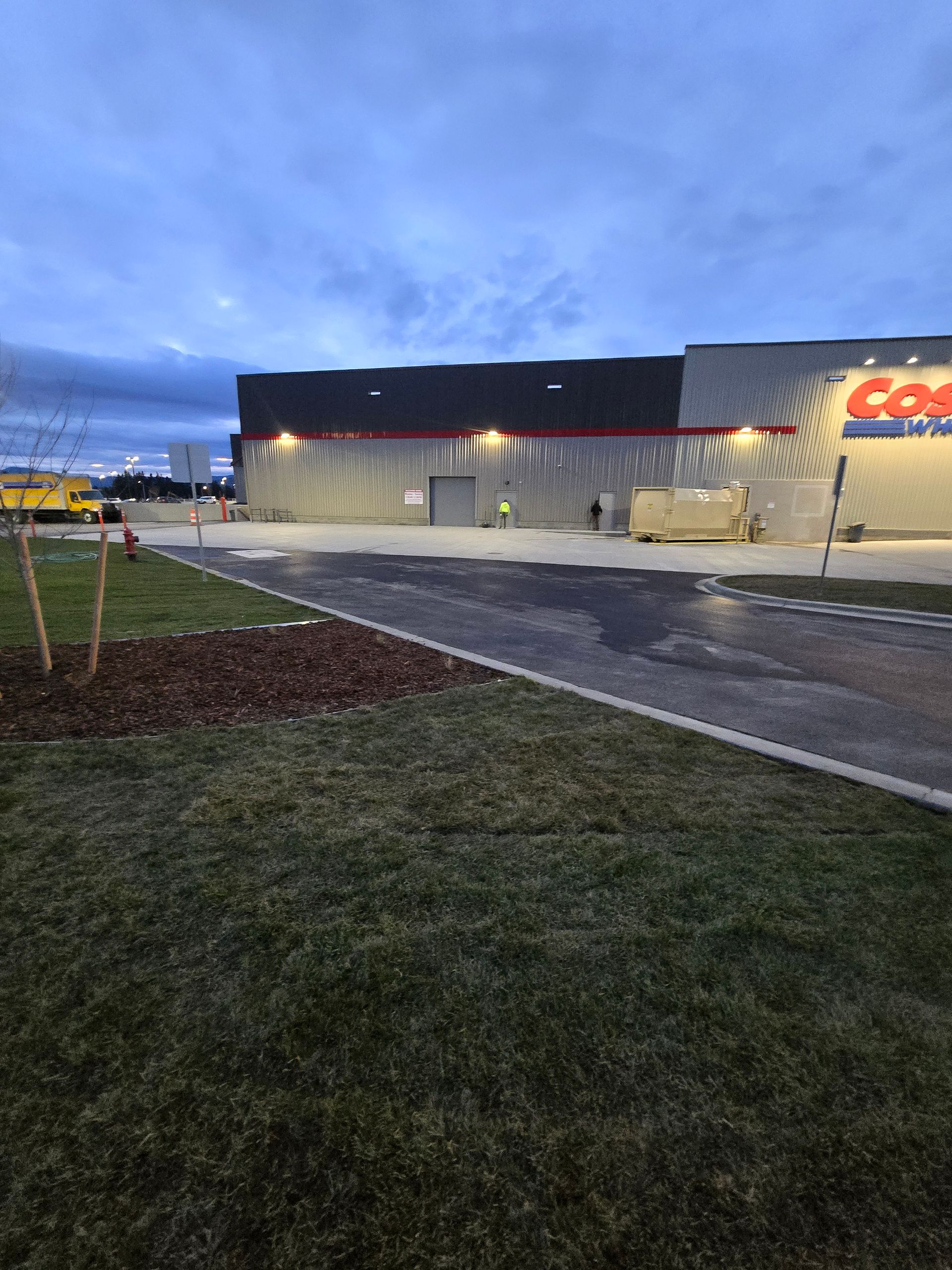 An exterior view of a Costco warehouse at dusk, featuring a large parking lot, grassy foreground, and bright wall lighting.