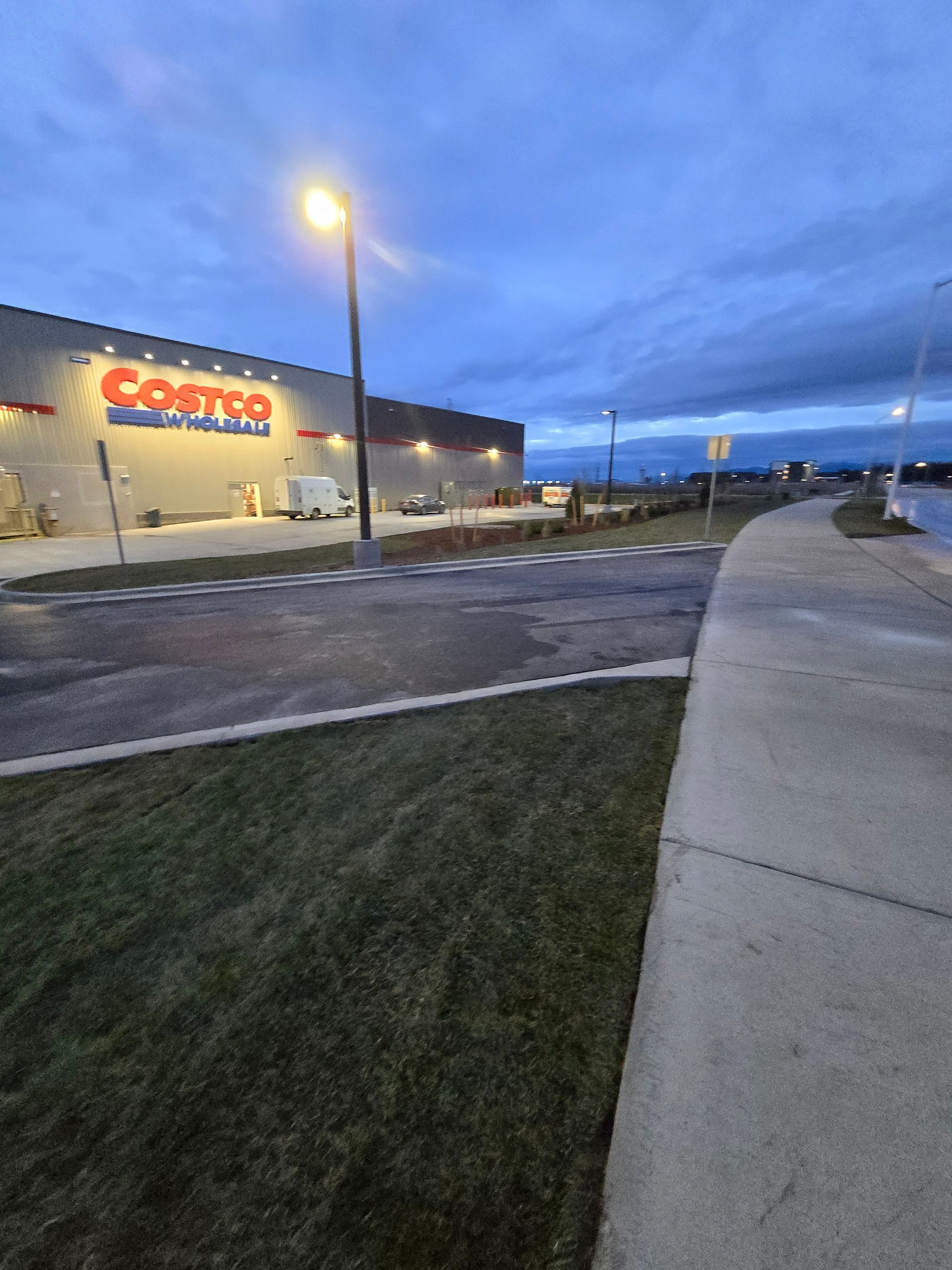 A Costco warehouse exterior at dusk, featuring a lit parking lot lamp, a grassy verge, and a concrete sidewalk.