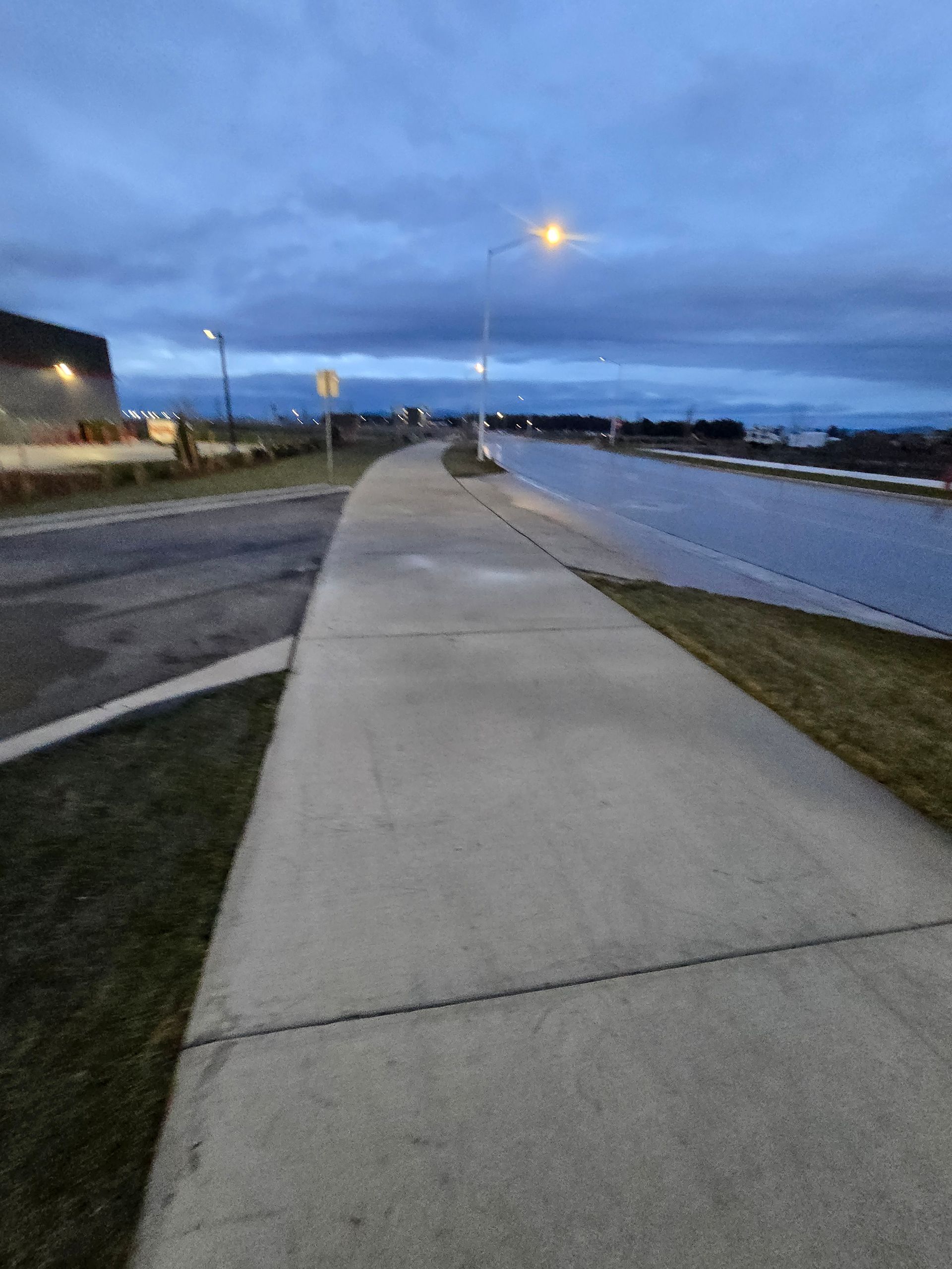 A paved sidewalk stretches into the distance at dusk, flanked by a grassy verge, a road, and a building under a cloudy sky.