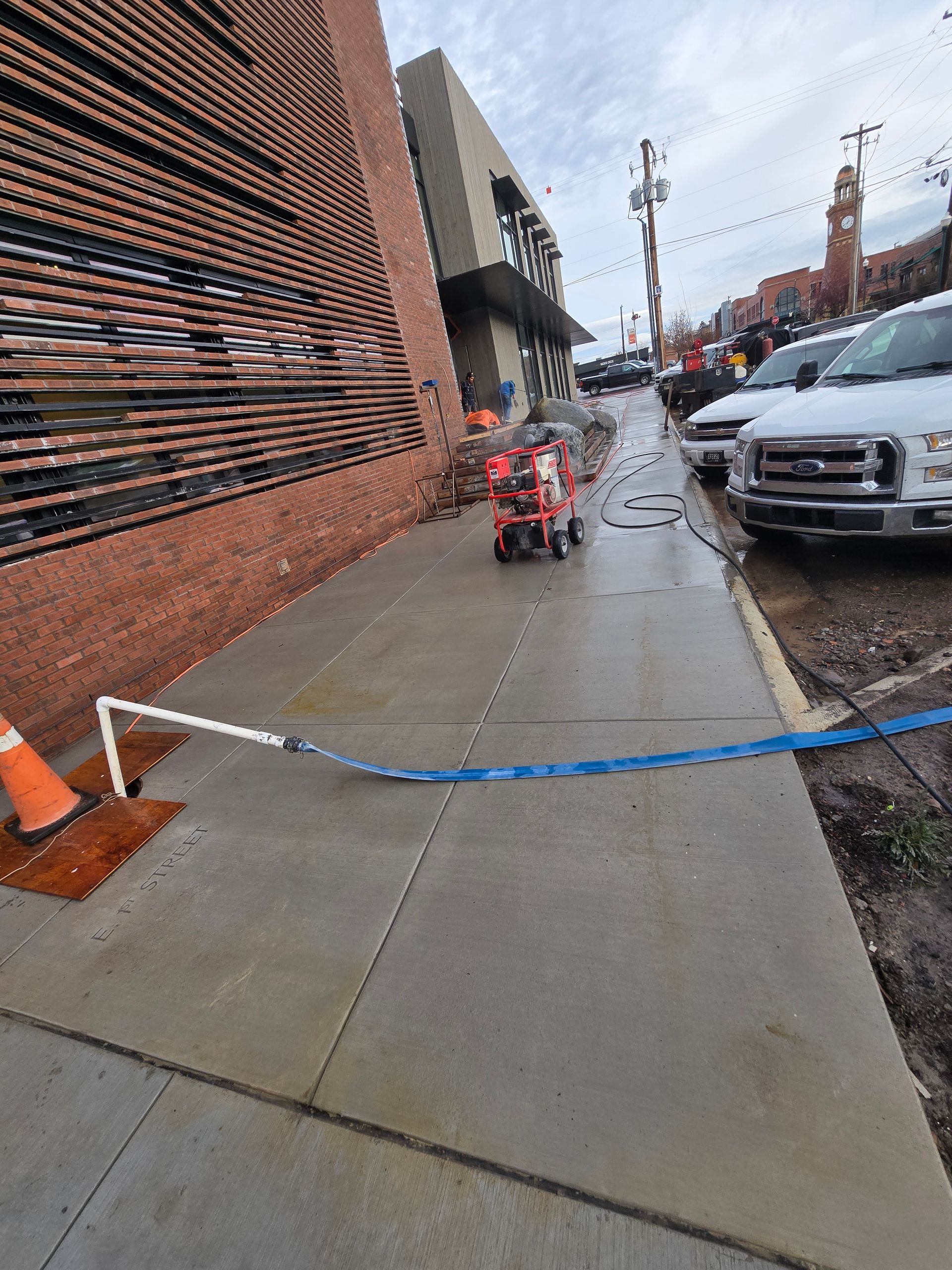 An orange traffic cone and blue tape barrier block a wet sidewalk next to a brick wall; a small red utility cart is ahead.