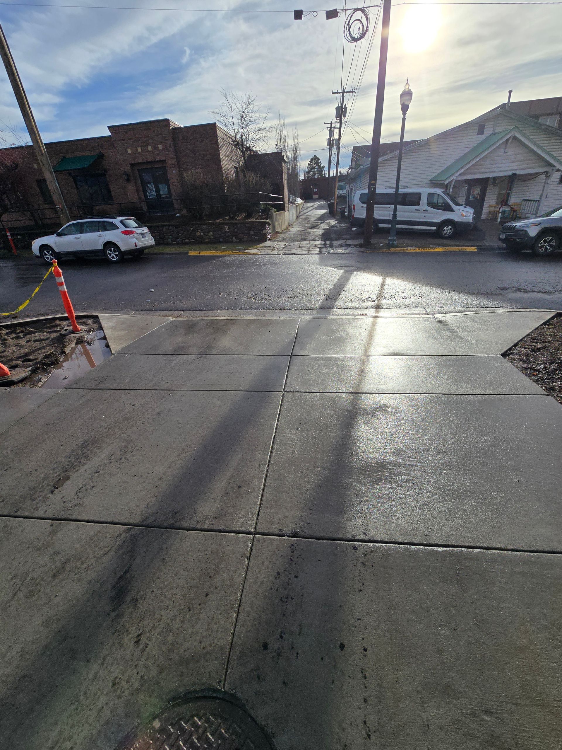 A sunny, urban street scene showing a sidewalk leading to an intersection with vehicles parked on the side.