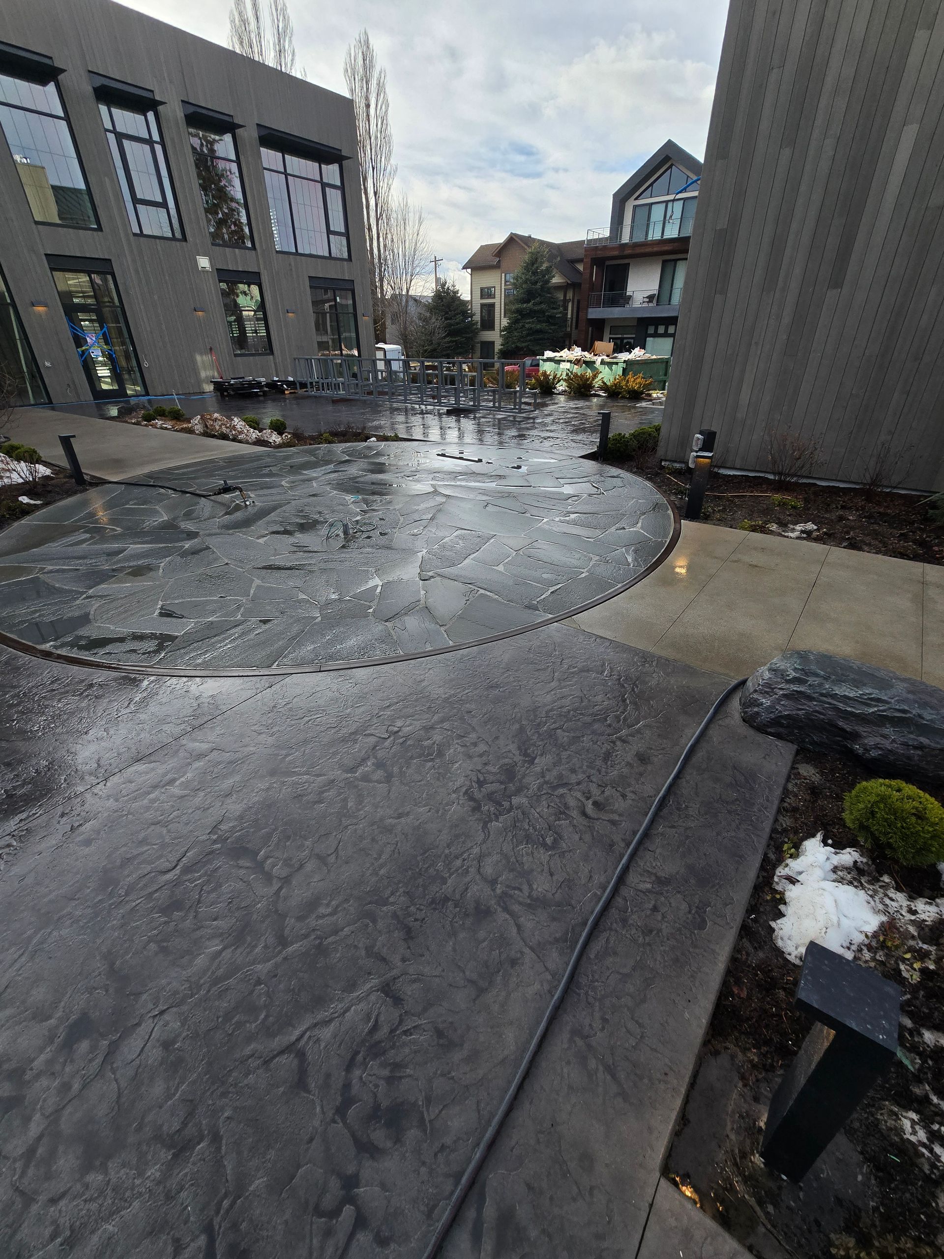 A gray, wet outdoor plaza between modern buildings, featuring a circular pattern on the ground and scattered stones.