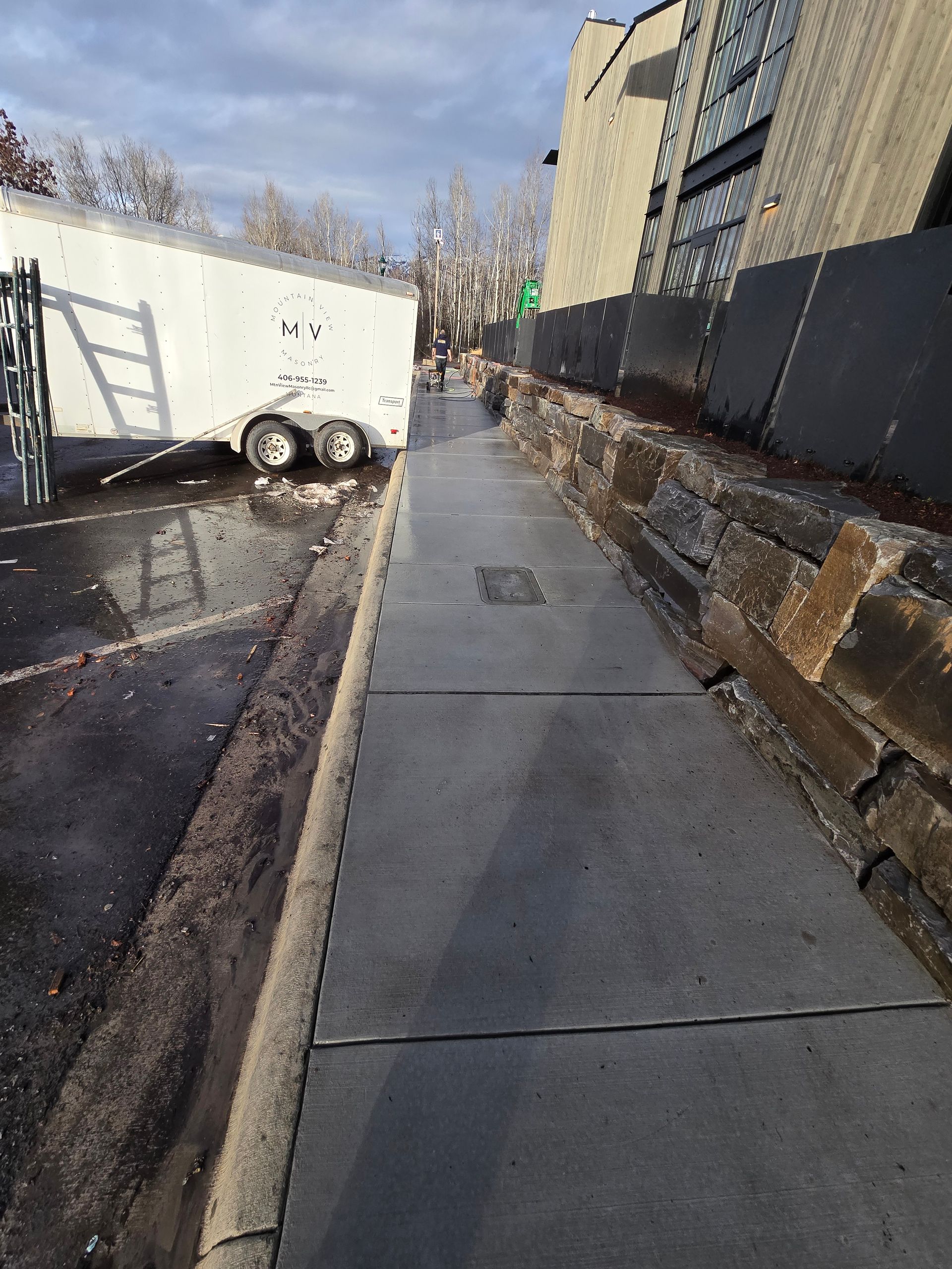 A paved sidewalk runs alongside a stone retaining wall and a white utility trailer parked on an asphalt lot.