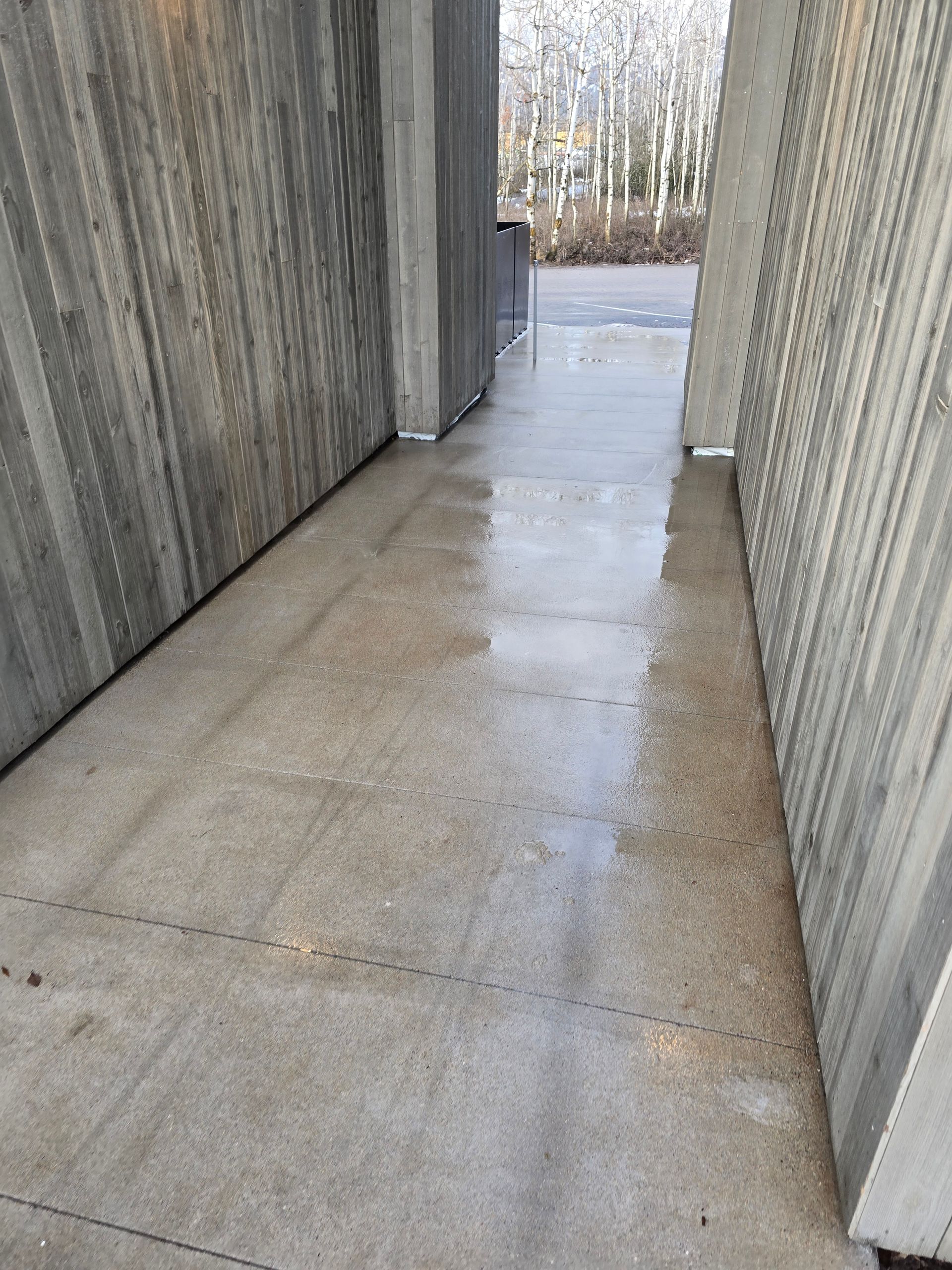 A wet concrete walkway leads between two textured concrete walls toward an outdoor area with trees in the background.