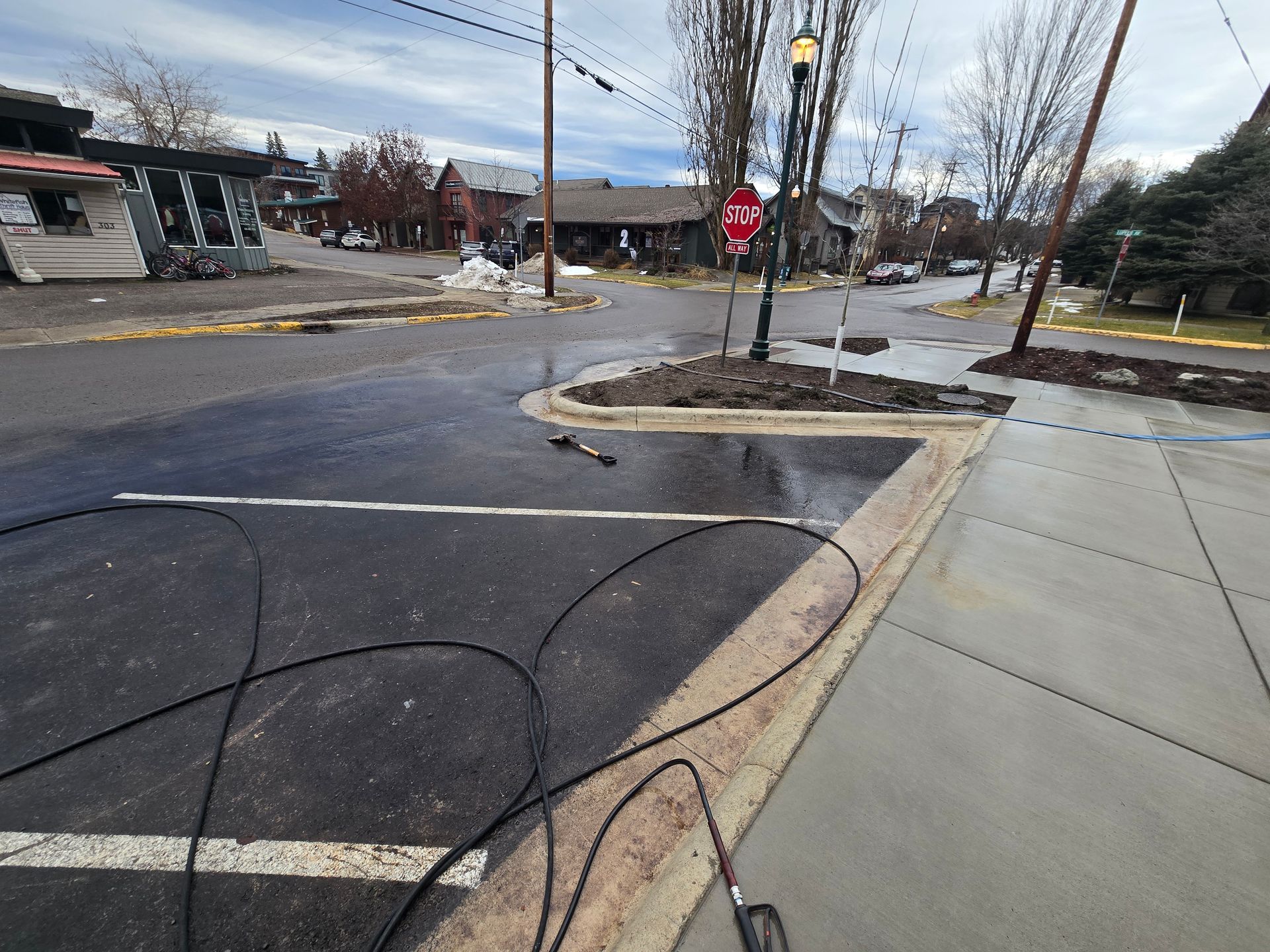 A freshly paved asphalt corner parking area with white painted lines, adjacent to a concrete sidewalk and landscaping.