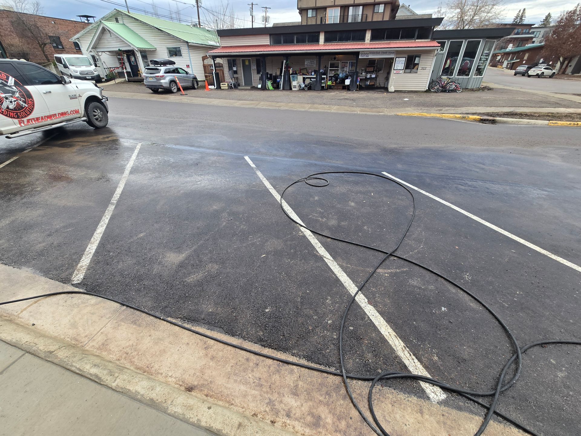 A wet, newly sealed parking lot with painted white diagonal lines and a black hose coiled on the dark asphalt.