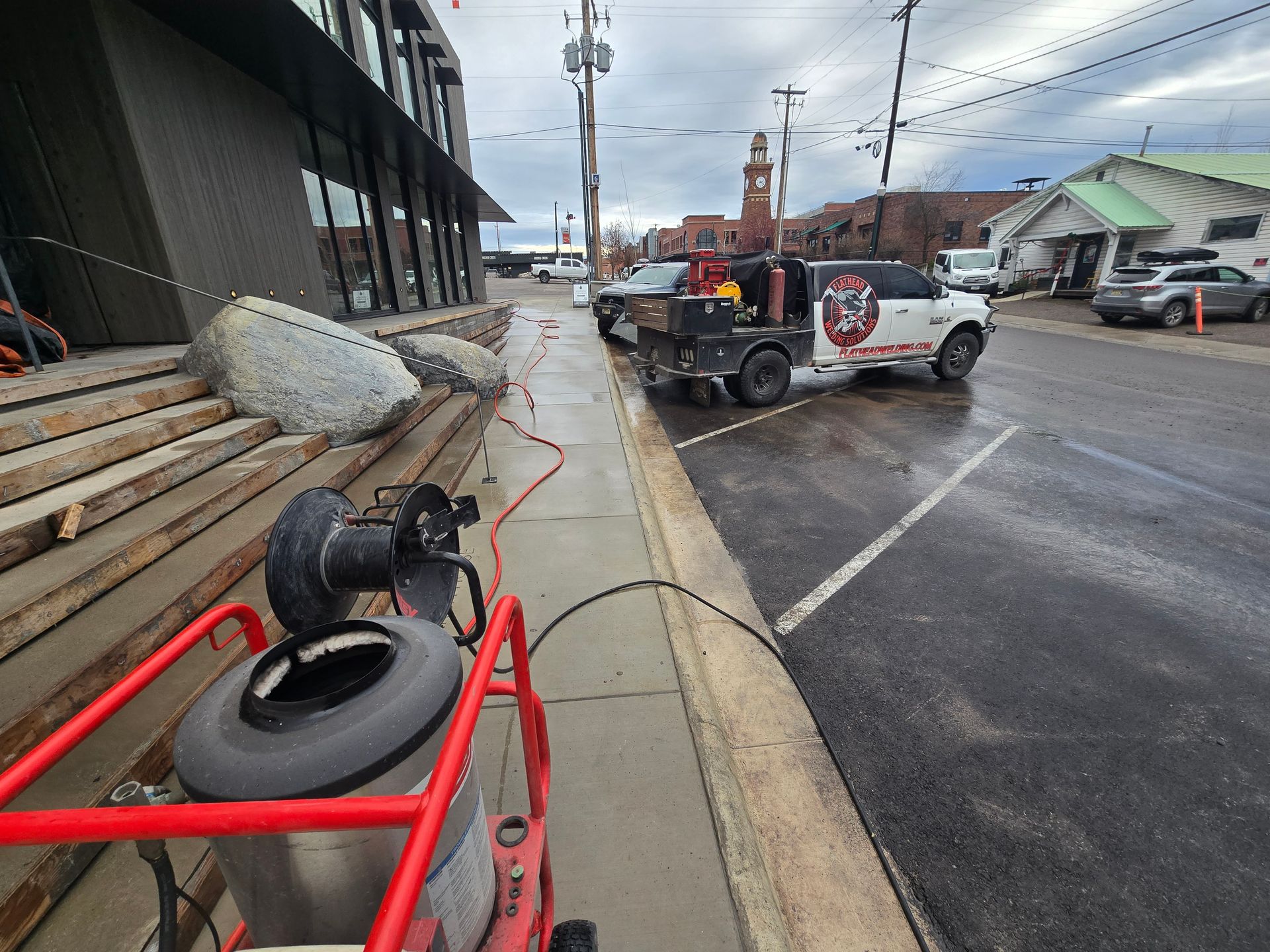 A red construction machine on a sidewalk in front of a building, with a work truck parked on the street nearby.